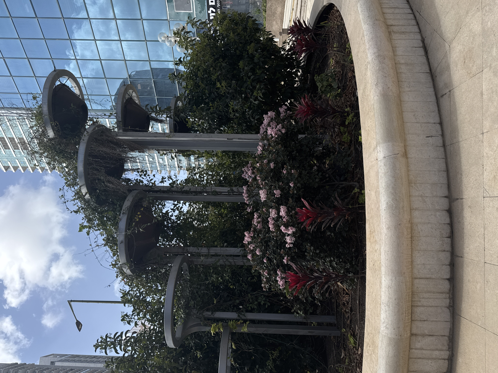 The image shows an outdoor scene in Ramat Gan, Israel. A three-tiered, circular planter is the main subject. Each tier supports plants and flowers, including bushes, creeping vines, and red-leafed plants. The planter structure consists of metal arches and supports. A light pole is visible in the background, as well as the reflection of buildings in a glass-covered structure. The sky is partly cloudy. The planter is made of light-colored stone. The photo seems to have been taken during the day.