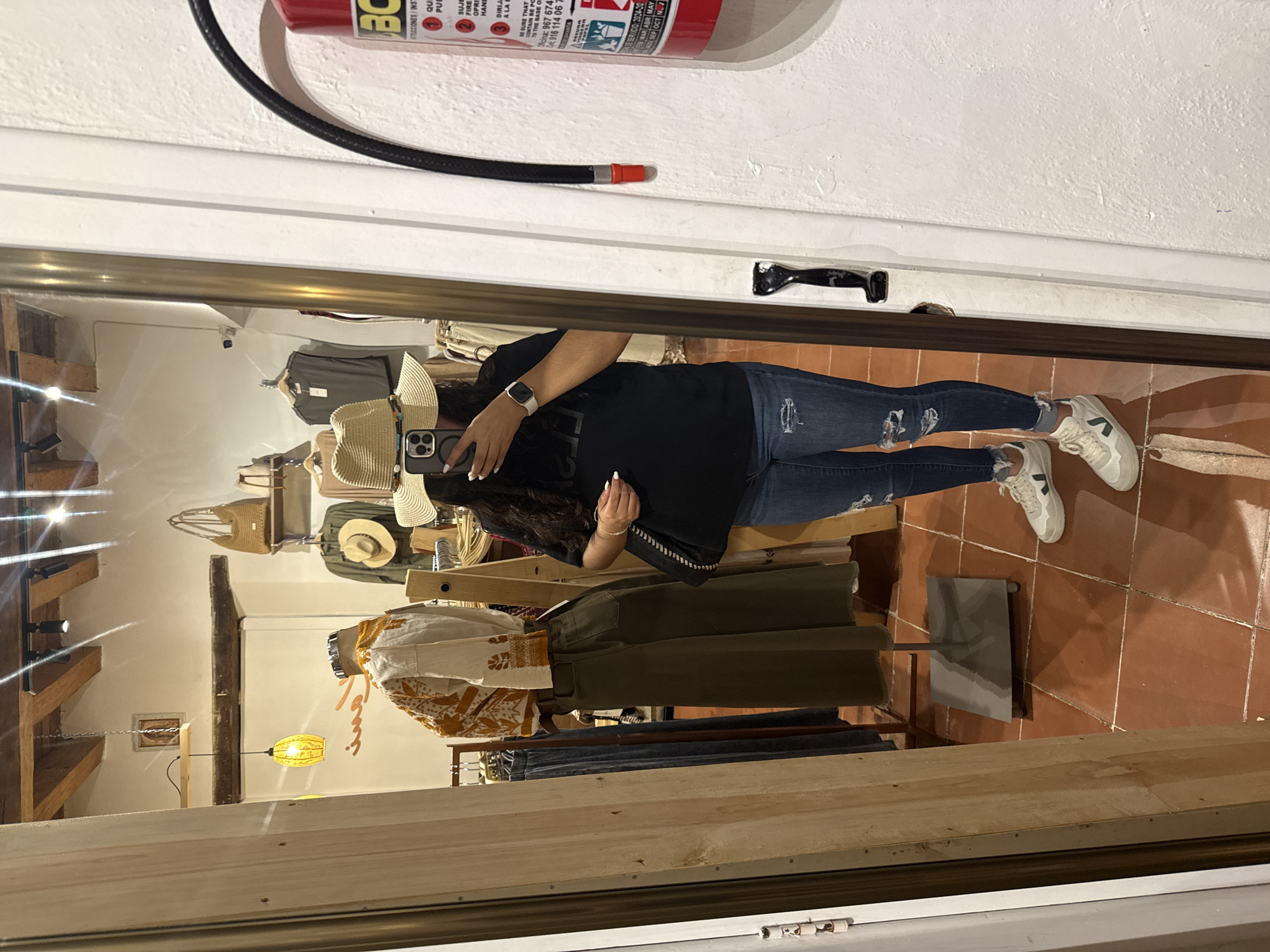 A reflection in a mirror shows a person taking a picture of themselves in a clothing store in San Cristóbal, Mexico. The person is wearing a black t-shirt, jeans with rips, and white sneakers. They are wearing a straw hat and an Apple Watch. The store has clothing racks and various articles of clothing, including tops, pants, and hats. A fire extinguisher is mounted above the mirror frame. The floor is tiled. A single pendant light fixture hangs in the store.