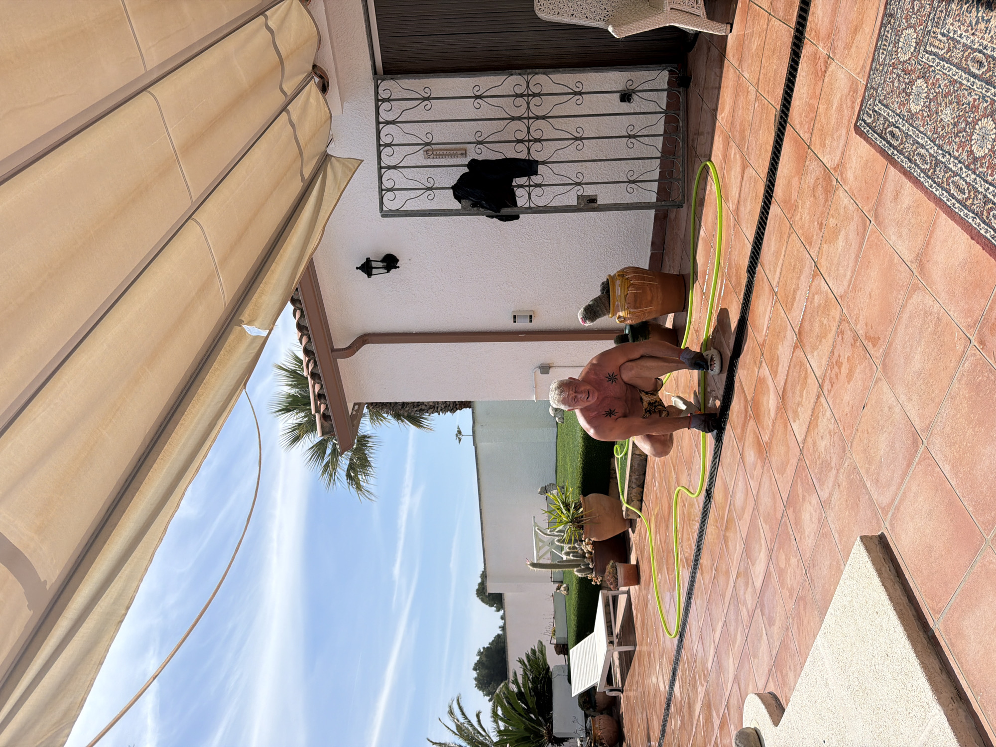 The image shows an outdoor scene in Mont-roig del Camp, Spain. A shirtless man with visible tattoos is crouching beside a green garden hose on a tiled patio. The patio features a drainage grate running along its length, with potted plants and a white patio set visible in the background. A building is behind the man and plants. The building's exterior has a white wall and a brown-tiled roof overhang. An awning is visible above the patio and sky and trees are visible in the background. A black jacket is hanging on the wall by the door of the house.
