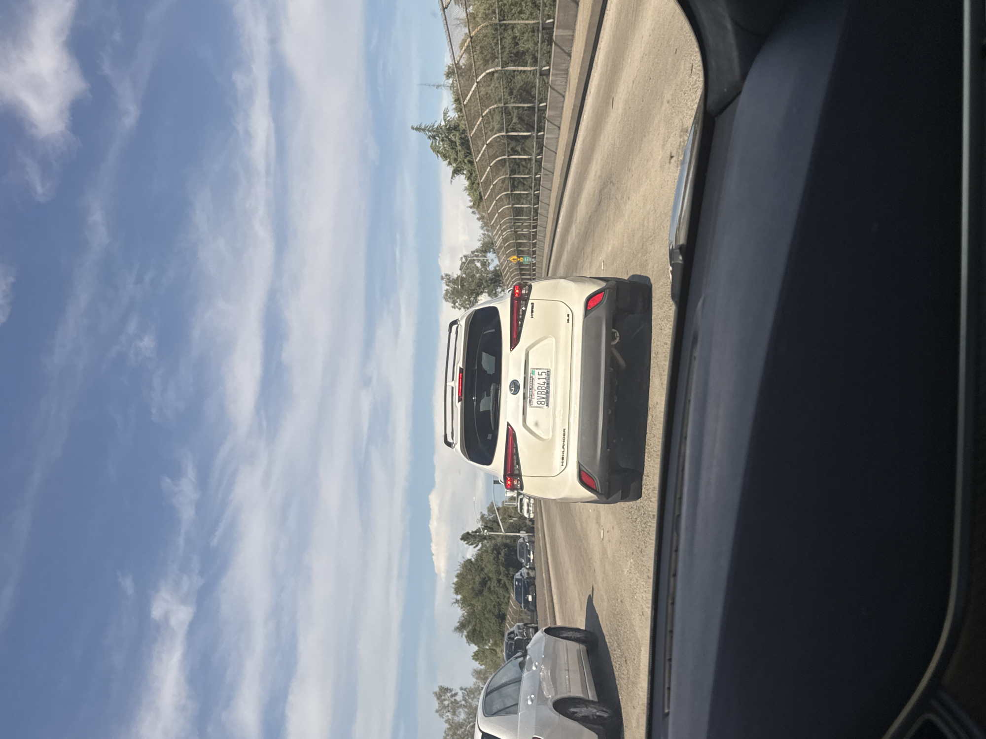 The image shows a view from inside a vehicle on a highway, likely in Citrus Heights, California, as indicated by the license plate frame on the white SUV. The primary focus is the rear of a white Toyota Highlander hybrid SUV with a roof rack, driving in the middle lane of a multi-lane highway. The license plate reads "8VBB415". To the left of the SUV, a white sedan is visible in the left lane. Further ahead, several other vehicles are present on the highway. The road is paved, with clear lane markings. On the right side of the highway, there is a concrete barrier with a chain-link fence on top, separating the road from vegetation and trees. The sky is blue with scattered white clouds. The overall scene depicts a typical traffic situation on a sunny day.
