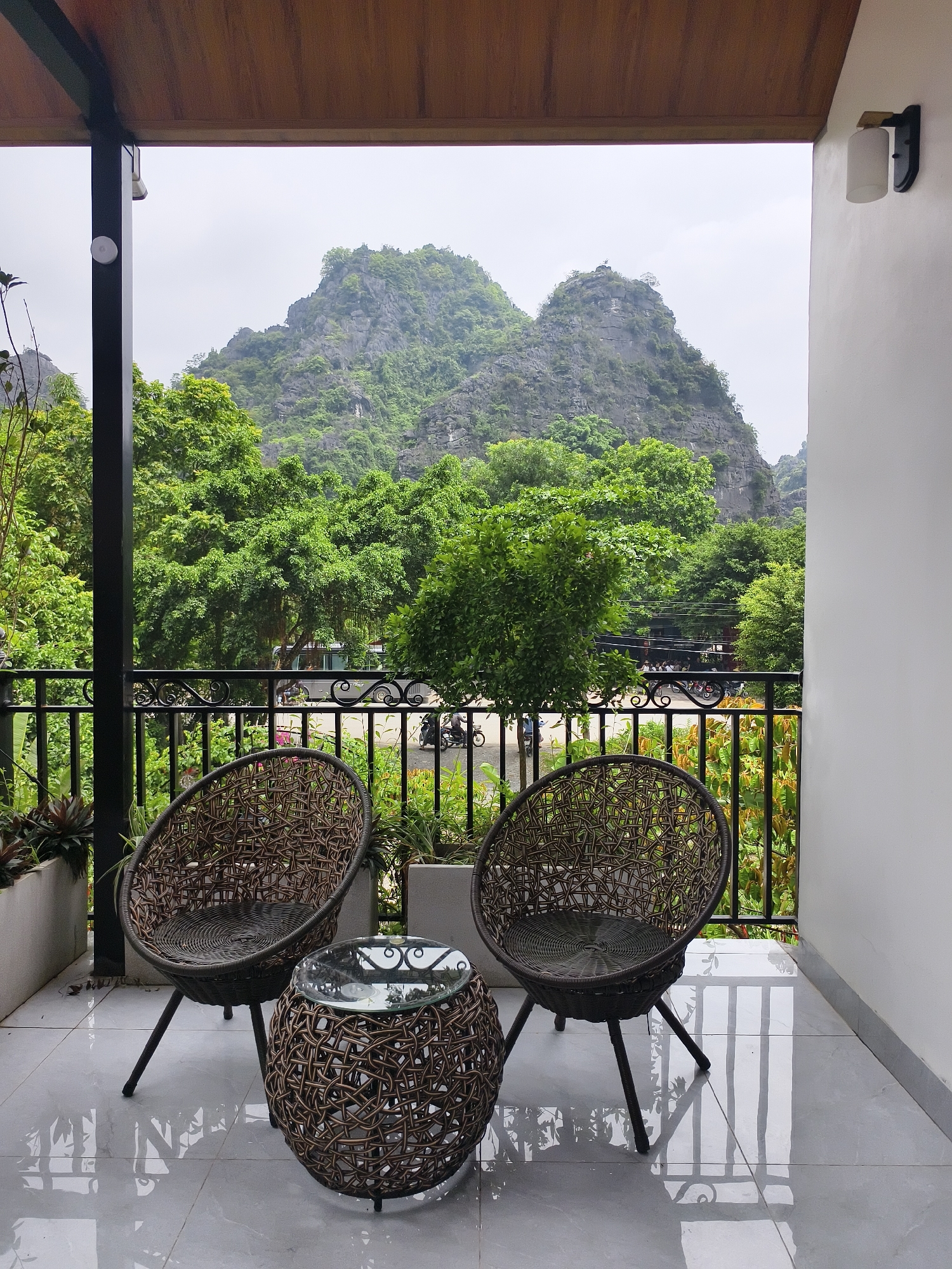 This image captures a balcony with a stunning view of karst mountains and lush greenery in Tây Hoa Lư Ward, Vietnam. Two rattan chairs and a matching coffee table with a glass top are arranged on a tiled floor. A black metal railing separates the balcony from the outdoors, beyond which a street is visible with some motorbikes and a bus. The background is dominated by a majestic, tree-covered mountain under an overcast sky. The overall scene suggests a tranquil setting for relaxation and enjoying the natural landscape.