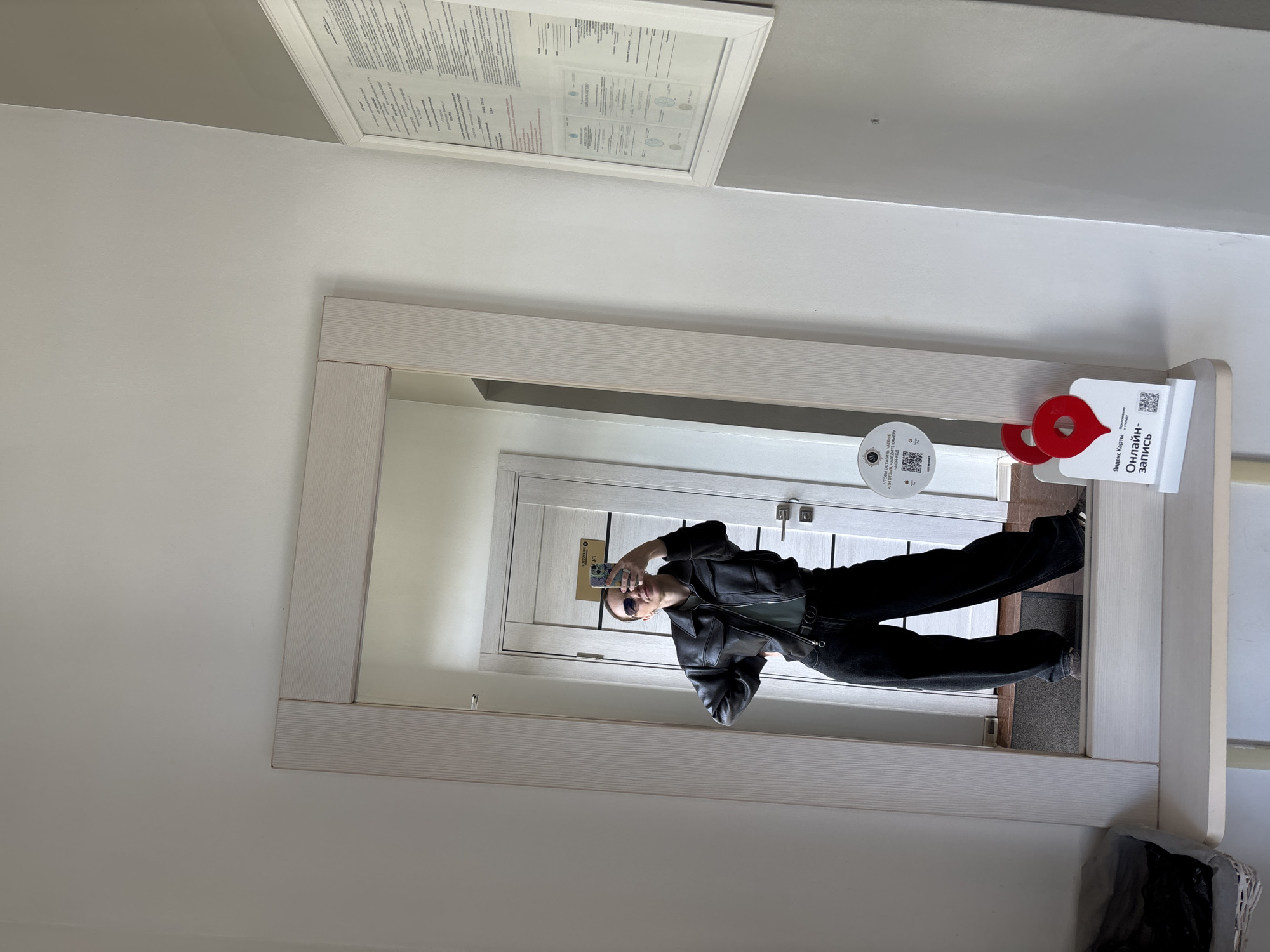 A person is reflected in a mirror taking a selfie. The person is wearing a black jacket, a green shirt, black pants, and sunglasses. The person is holding a cell phone in their left hand to take the picture. The mirror is rectangular with a light wood frame. The mirror is hanging on a white wall. Above the mirror is a document, which is also framed. To the right of the mirror is an online booking tag. The reflection in the mirror shows a white door with a silver handle. The location is in the city of Unknown, Unknown.