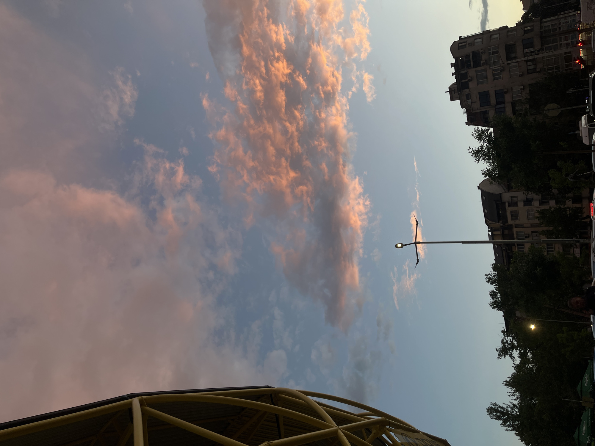 The image captures a street view in Plovdiv, Bulgaria, at what appears to be dusk. The sky is predominantly light blue, adorned with streaky clouds tinted with orange and pink hues, likely from the setting sun. Below, the corner of a yellow metal structure with a curved design is visible. A lamppost extends into the sky, its light not yet activated. The backdrop includes multi-story buildings with visible windows and balconies, partially obscured by trees. Cars are also parked on the streets with trees.