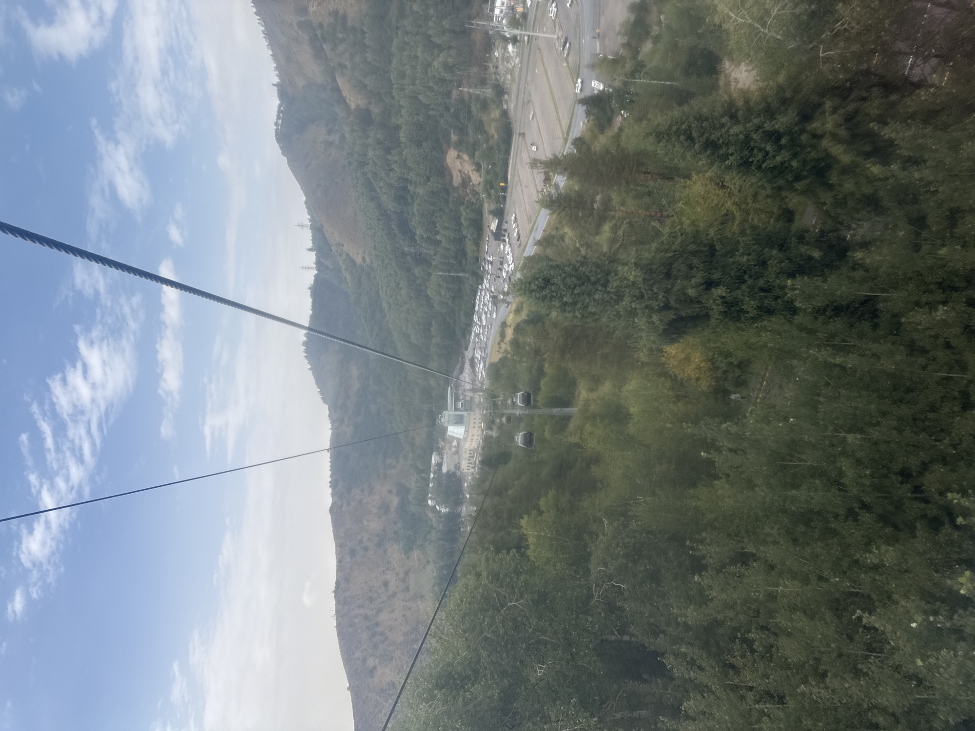 The image captures a view from a cable car in a mountainous, forested area with a city or large complex visible in the distance. The cable car's rope is prominently featured in the foreground, running diagonally across the frame. Below, a dense canopy of green trees covers the slopes of a hill. In the mid-ground, a building complex with light-colored structures and a distinct glass-paneled roof is situated near a large, paved area filled with numerous parked cars. Further into the distance, the terrain continues with more hills and trees, and power lines can be seen on the higher slopes under a hazy, pale blue sky with scattered white clouds. Two cable car cabins are visible suspended from a cable, moving towards the distant buildings.