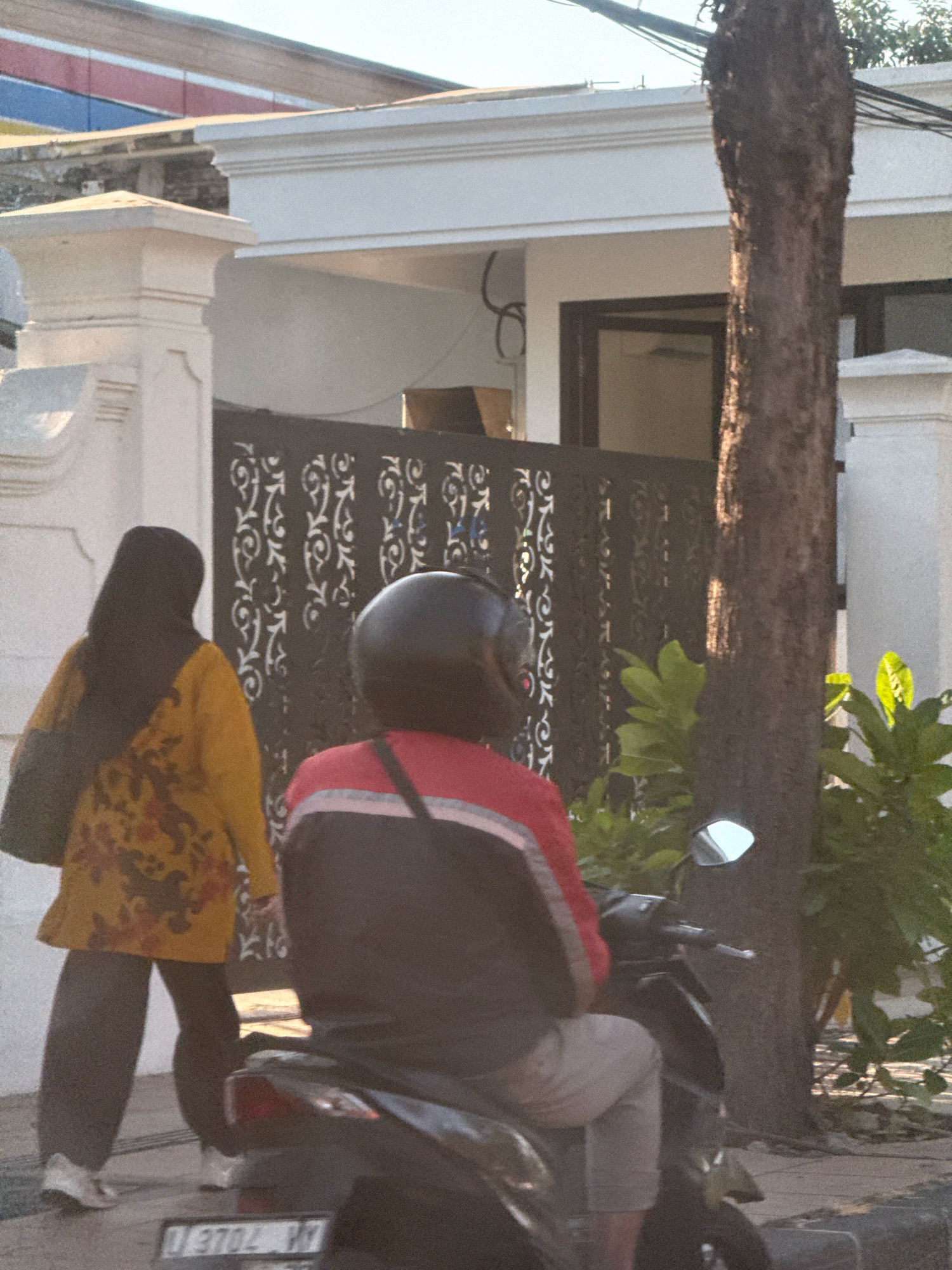 A medium shot shows two individuals on a street, with a motorcycle in the foreground. The person in the foreground, viewed from behind, is wearing a red and black jacket and grey pants, riding a black motorcycle. To the left, a person from behind wears a yellow tunic with a floral pattern and a black hijab, dark pants, and white shoes, carrying a black bag. The background reveals a white building with decorative fencing and a large tree trunk. The scene appears to be in a residential area, possibly in Surabaya, Indonesia, indicated by the presence of a vehicle license plate with Indonesian characters. The lighting suggests it is daytime, with shadows cast by the tree.