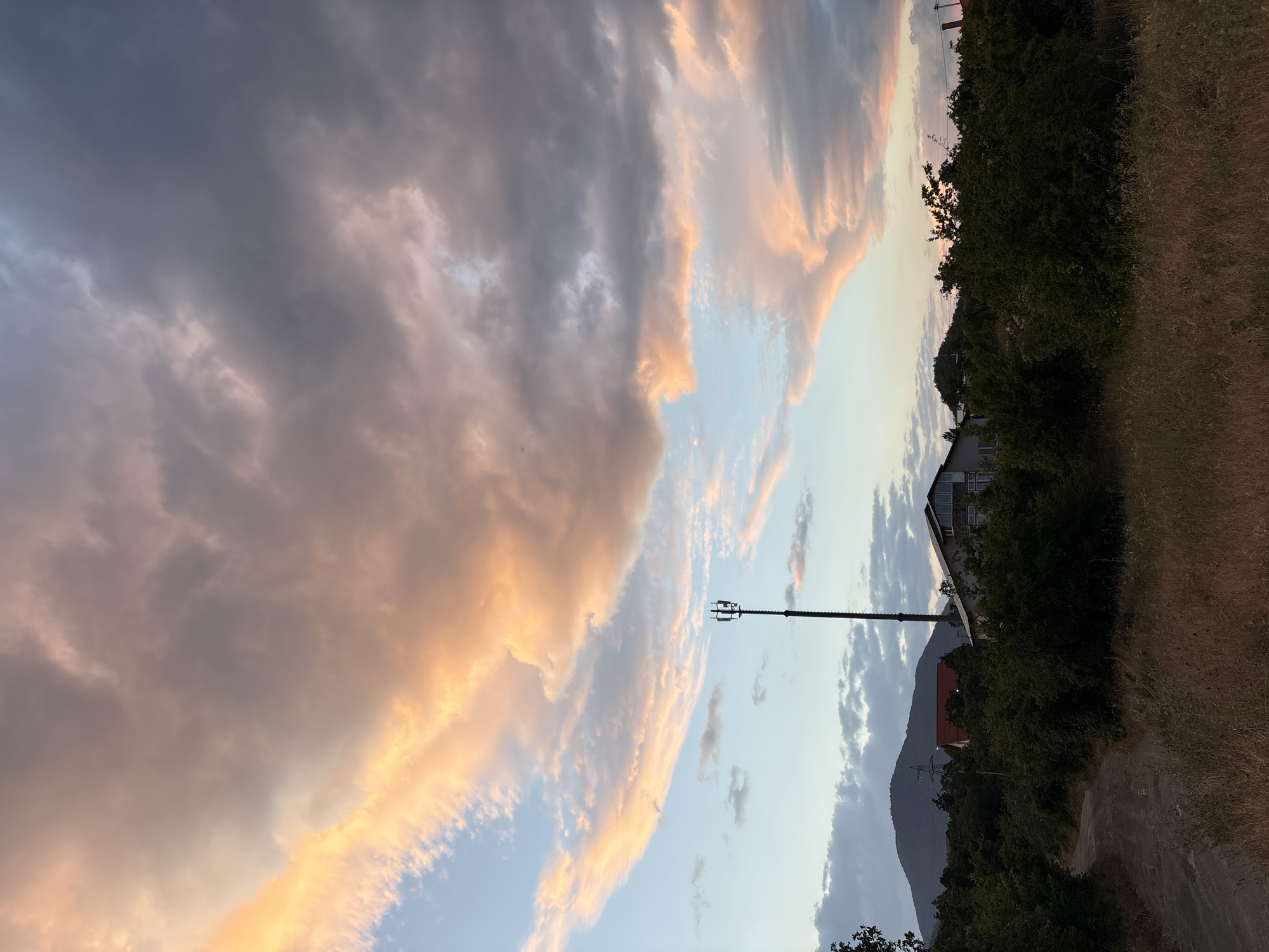 This image depicts a twilight sky over a rural area, likely in Malyi Mayak, Ukraine. The sky is filled with dramatic clouds, illuminated with hues of orange, pink, and grey, suggesting the setting sun. In the mid-ground, a tall, slender communication tower with antennas stands prominently. Adjacent to it are several buildings, including a house with a red roof and another larger structure with multiple windows. The foreground features a dirt path winding through dry grass and surrounding green foliage. In the background, a silhouetted mountain range can be seen under the cloudy sky.