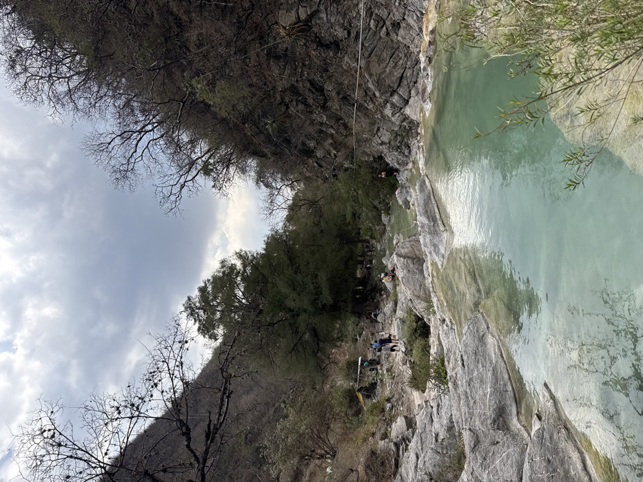 The image displays a valley scene in San Miguel Atlapulco, Mexico, under an overcast sky with scattered grey and white clouds. A natural waterway, characterized by multiple pools of clear, light turquoise-green water, flows through the center foreground and middle ground, bounded by large, smooth, grey-white rock formations.
Steep, rocky hillsides flank both sides of the valley. The hillsides are covered with a mix of vegetation, including numerous deciduous trees with bare branches, particularly on the upper slopes, and denser evergreen foliage closer to the valley floor. Sparse shrubs and grasses grow among the rocks and along the water's edge. On the right side, a dark, rocky cliff face rises steeply, displaying some small agave-like plants.
Approximately 10 to 15 individuals are present within the scene. On the left bank, in the middle distance, a dirt path leads towards a rudimentary shelter with a yellowish-tan tarp roof. A group of approximately four individuals are walking on this path near the shelter. Other individuals are dispersed along the rocky banks of the waterway, standing or sitting. One individual in dark clothing is visible near the water's edge against the rocky cliff on the far right. A thin, light-colored rope or cable stretches horizontally across the upper middle ground, attached to the right cliff face.
