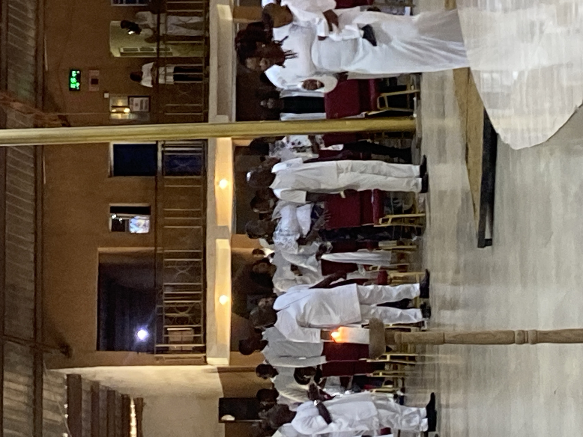 The image shows a group of people in a church, all dressed in white, standing in rows. They appear to be participating in a religious ceremony or service. The location is Rukuba Road, Nigeria. The interior is illuminated, and the scene includes wooden pews and other religious artifacts.