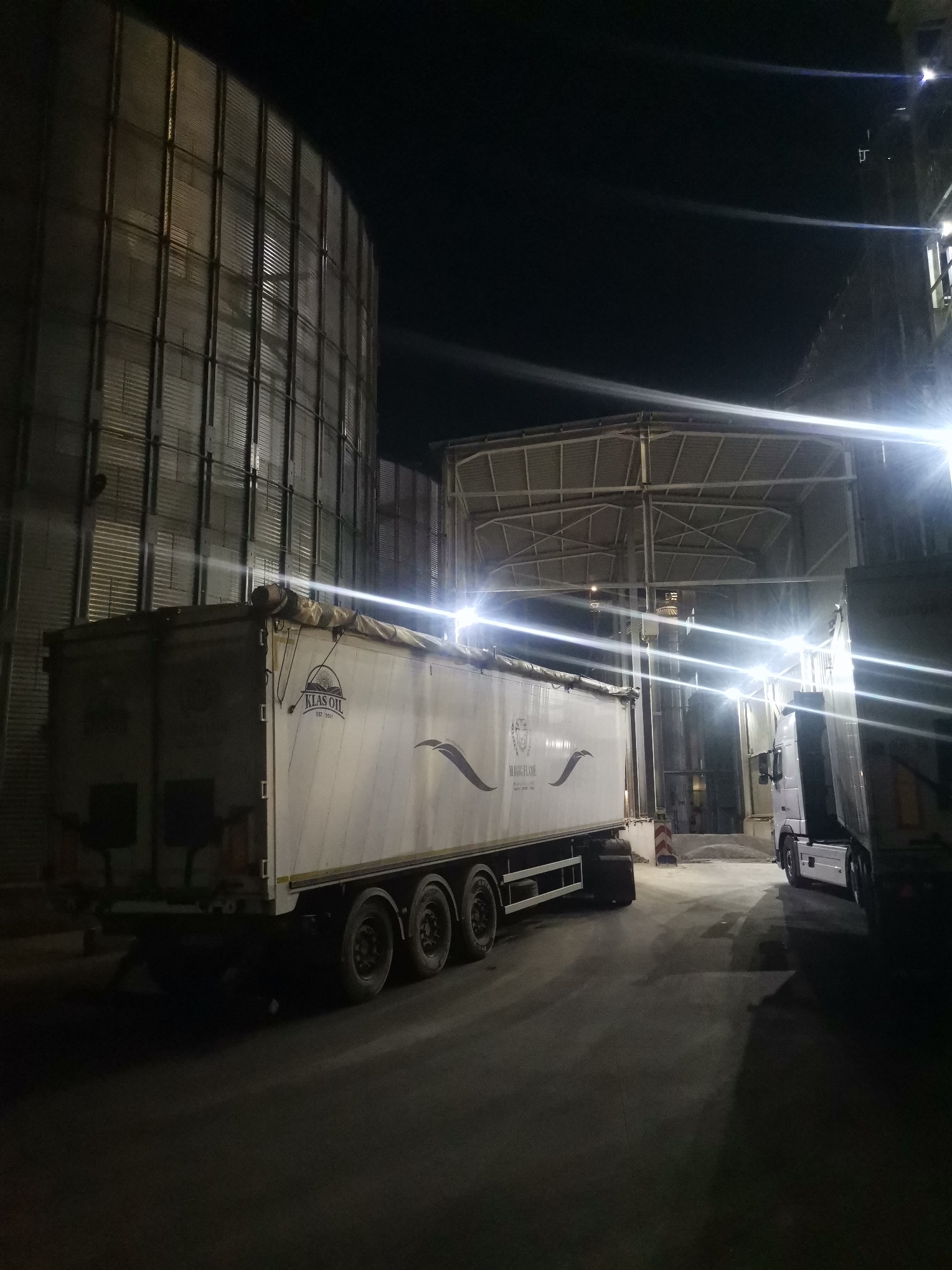 At night in Razdelna, Bulgaria, a white semi-trailer with "KLAS OIL EST. 2001" printed on the side sits beside a large metal grain silo. Another truck is parked nearby under a metal framework structure with bright lights. The ground is asphalt.