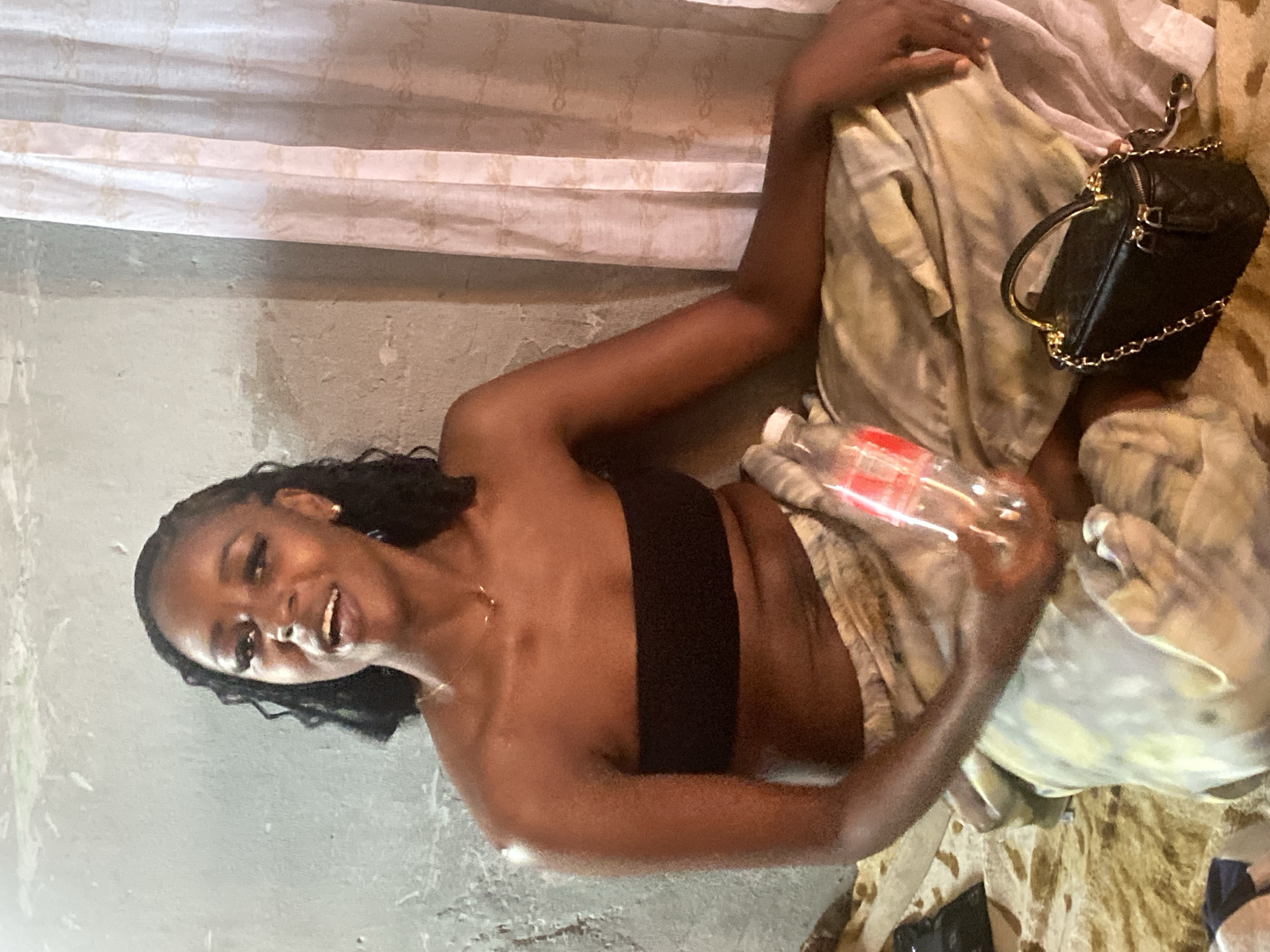The image, taken in Sabon Tasha, Nigeria, depicts a young Black woman seated indoors, looking directly at the camera with a broad smile and her mouth slightly open, suggesting she is speaking or laughing. She has dark, curly hair and is wearing a black strapless top, along with a delicate gold chain necklace.
She is positioned on or wrapped in a light-colored, patterned fabric, possibly a blanket or clothing, which features shades of beige, green, and white. In her right hand, she holds a clear plastic bottle with an unreadable red label. Next to her, on the patterned fabric, lies a small, black quilted handbag with a gold chain strap.
The background consists of a light grey or beige concrete wall with a somewhat rough texture. Above the wall, a white or off-white curtain or drape is visible. The lighting appears to be artificial or from an indoor setting, creating a warm tone. The woman's expression conveys happiness and ease. No explicit time of day, weather conditions, or additional visible text are discernible from the image.