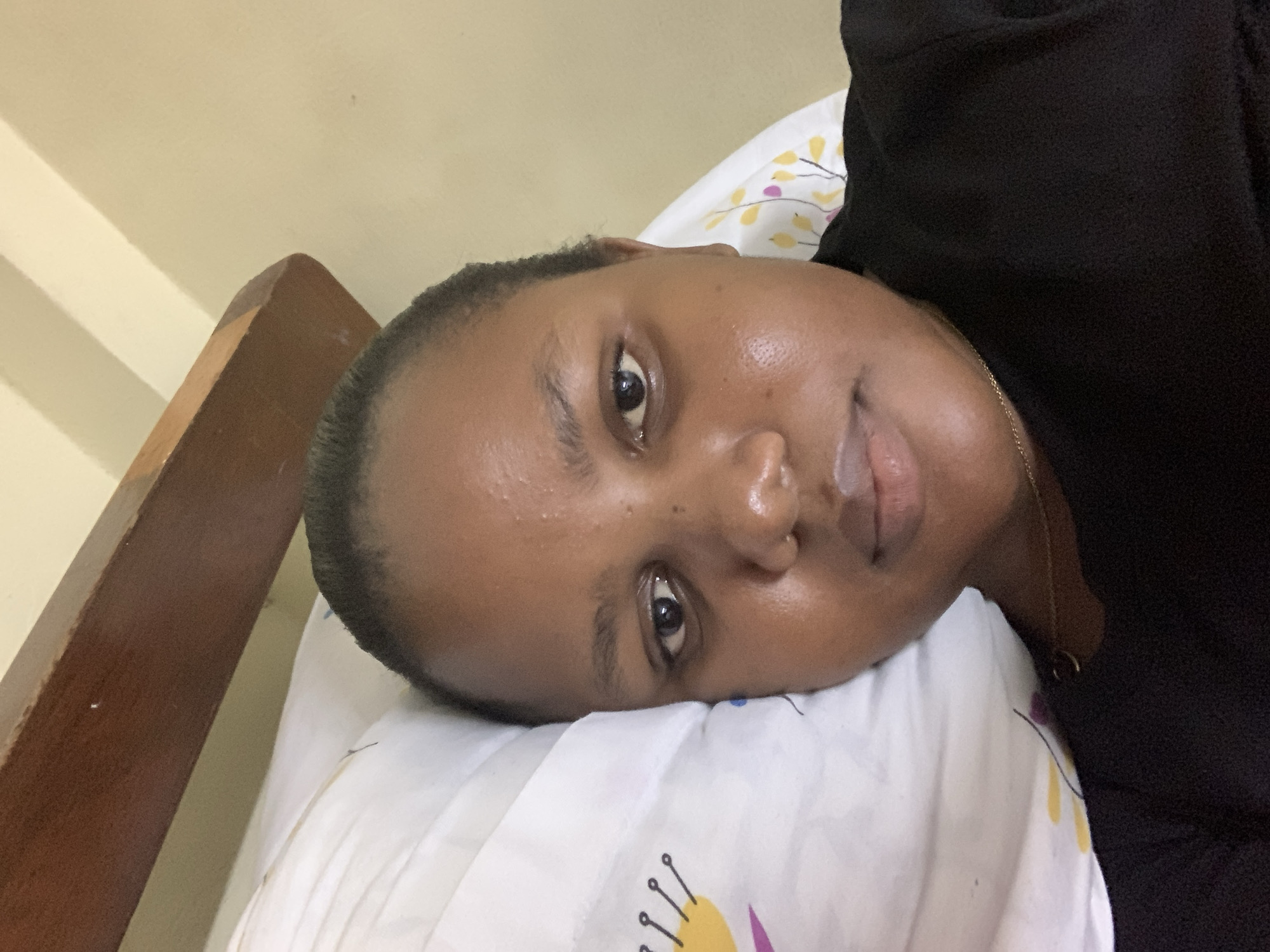 A medium shot depicts a woman lying on her side on a pillow with a patterned design, likely in a residential setting. Her head rests on the white pillow with yellow and purple floral-like patterns. She has dark hair, dark eyes, and dark skin. She's wearing a black top and a gold necklace is visible. The environment includes a beige wall and a wooden bed frame. There is no verifiable information connecting the image to Dar es-Salaam, Tanzania.