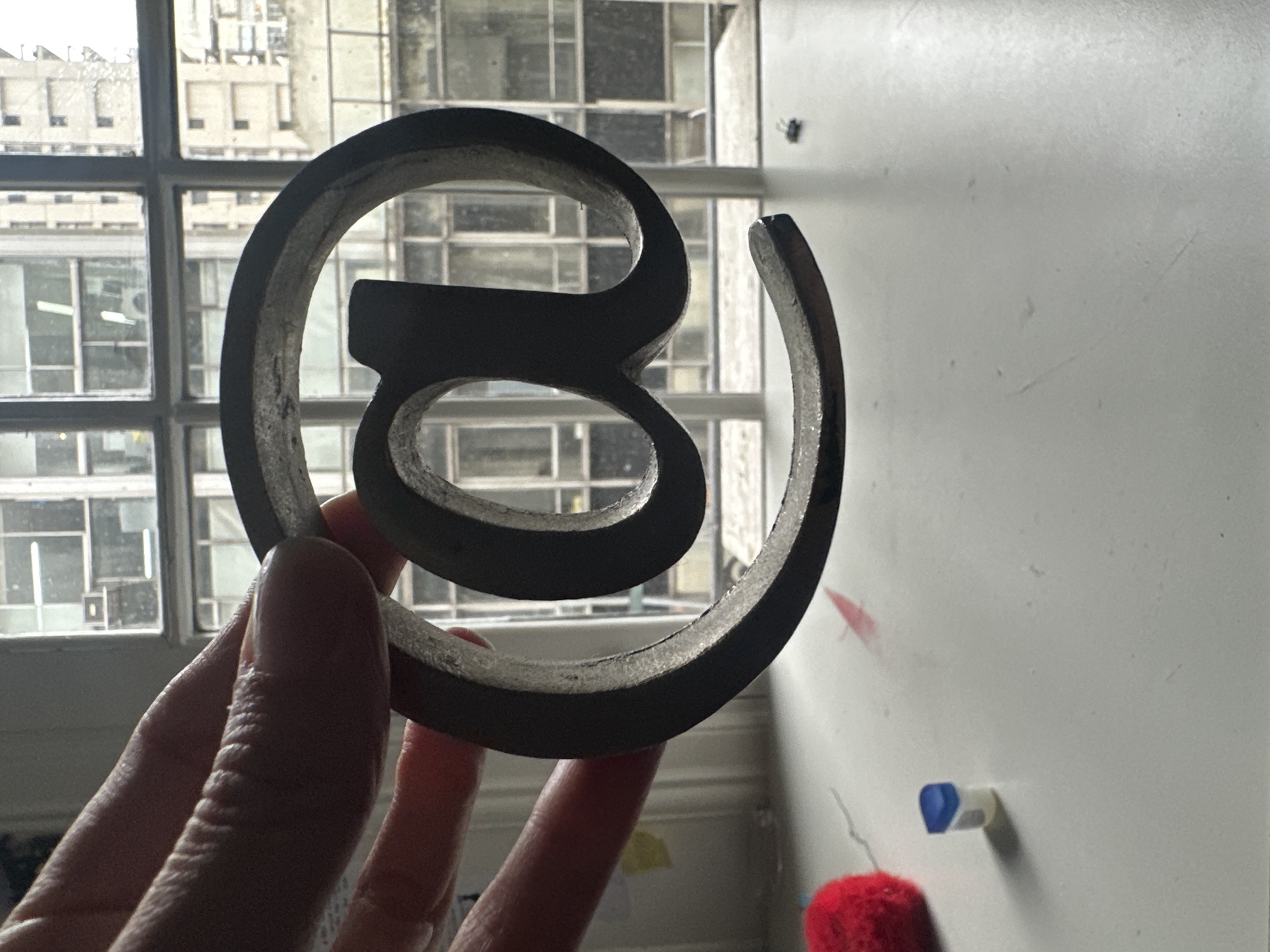 A person's hand holds a metal symbol resembling a stylized "@" sign in front of a window. The window reveals a cityscape with multiple buildings. The background also includes a white wall with a few smudges and objects. A blue-and-white eraser and a red fuzzy object are near the bottom of the wall. The image appears to be taken indoors, with natural light coming from the window. There is no information provided in the picture to determine the location of the city of Buenos Aires, Argentina.
