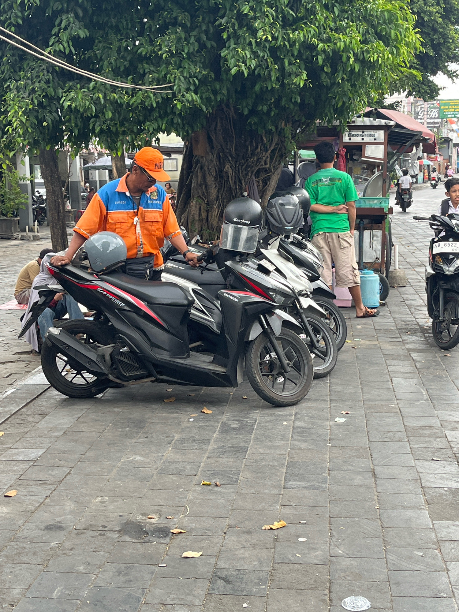 This is a medium shot photo of a man wearing an orange shirt with blue accents and a bright orange cap, standing next to several parked motorcycles on a cobblestone street in Yogyakarta, Indonesia. The man, who appears to be a parking attendant, has "SUHADI" and "DINDOPAS" printed on his shirt. He is looking down and to his right, with his left hand resting on the seat of a black motorcycle. The motorcycle has a gray helmet on its seat.
Behind the man, two more helmets are visible on the seats of other motorcycles. Further back, a person in a green shirt is standing with their arms crossed, and next to them is a food cart with a sign that reads "BAKSO RP 10.000". Several other people and motorcycles are visible in the background, suggesting a busy urban environment. A large tree with dense green foliage provides shade over the scene. The cobblestone street is littered with a few fallen leaves and small pieces of debris. The overall lighting suggests it is daytime, likely overcast or with dappled sunlight.