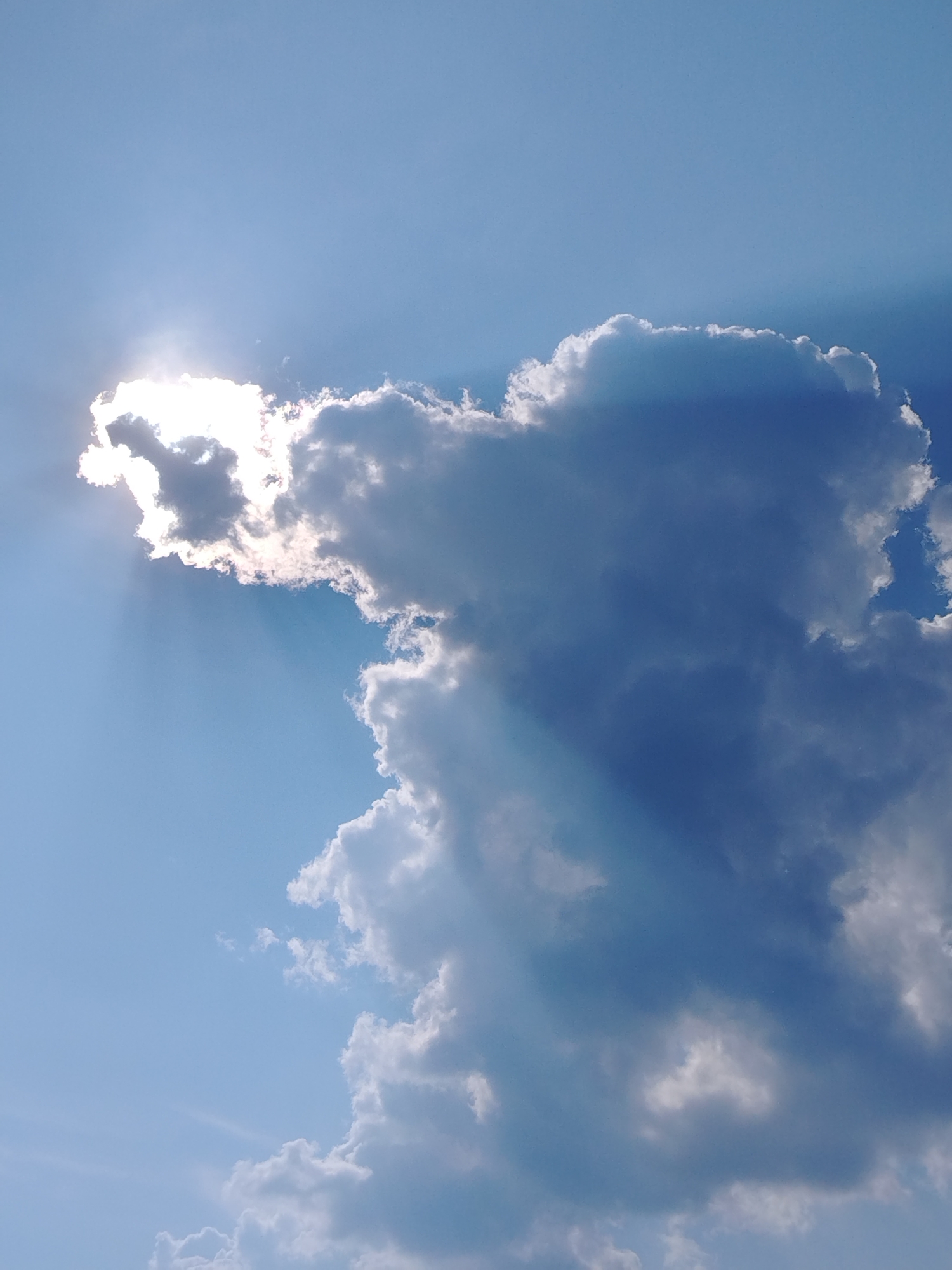 A bright sun is partially obscured by a fluffy white cloud, casting rays of light across a clear blue sky. The sun, located in the upper left quadrant of the image, creates a dramatic halo effect as its light filters through the edges of the cloud. The cloud itself is voluminous and has a textured, wispy appearance. A larger, darker cloud formation occupies the right side of the frame, with its shadow casting a blueish tint on the sky. The overall scene captures a moment of dynamic weather, with light and shadow playing across the expansive sky. The location is identified as Chaikovsky, Russia.