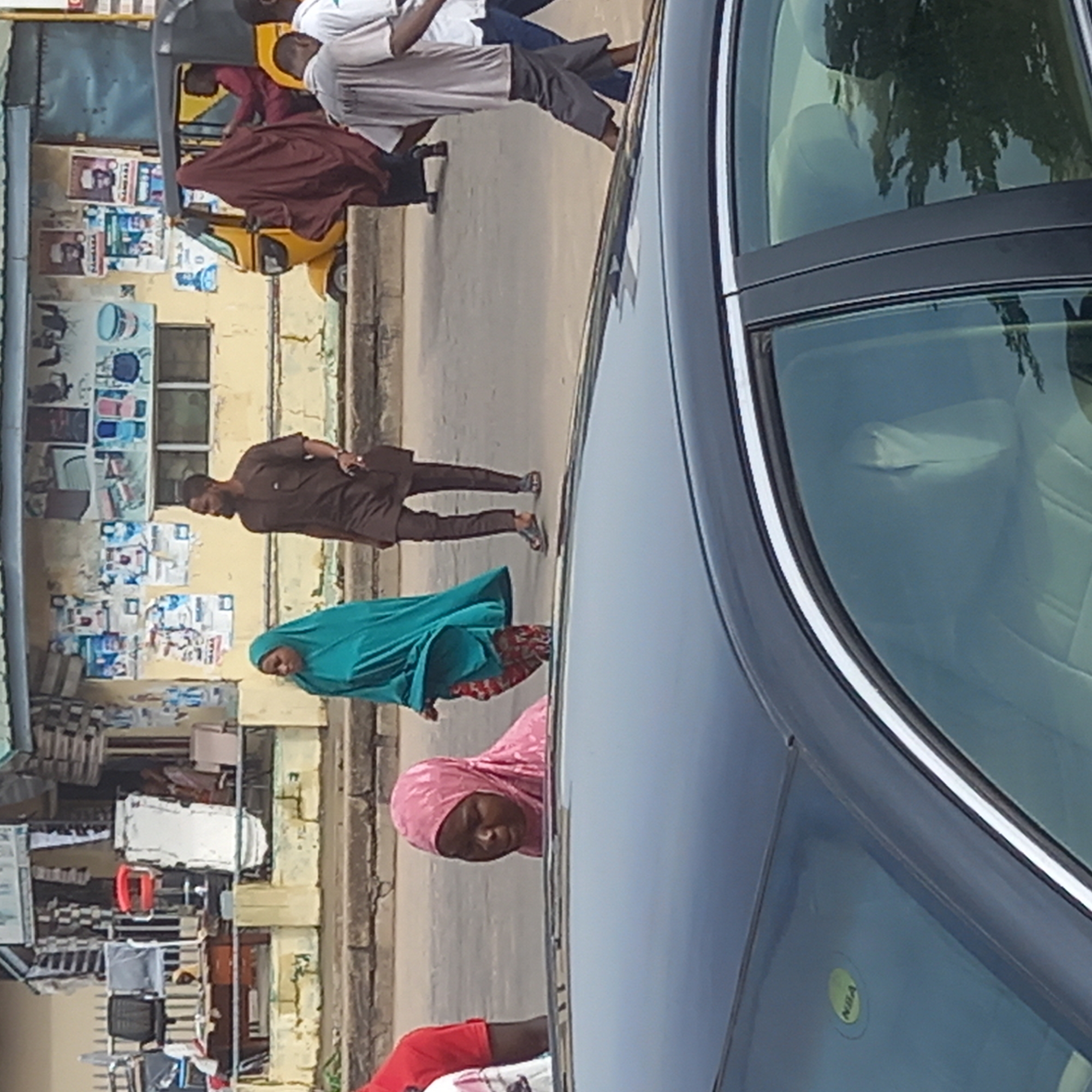 The image shows a street scene in Dadi Riba, Nigeria. A dark-colored car occupies the right side of the frame. On the street, several pedestrians are walking. One woman wears a pink headscarf, another a green headscarf, and a man is dressed in brown traditional attire. In the background, a yellow motorized tricycle ("Keke Napep") is visible near a building covered with posters. The storefront has shelves of goods on display.
