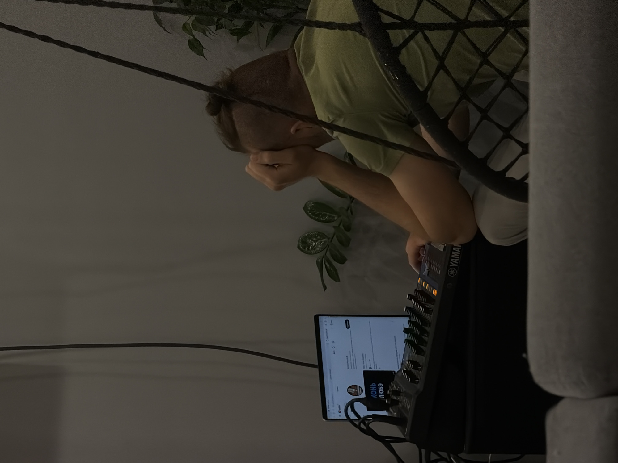 A man sits in a hammock, head in his hands, looking at a laptop and a Yamaha mixing console. The laptop displays a YouTube page with the channel "Конь Любэ" (Horse Lyube), which suggests the image was taken in Russia, potentially in the city of Chaikovsky, as this information was provided in the prompt's request. A houseplant is visible in the background.