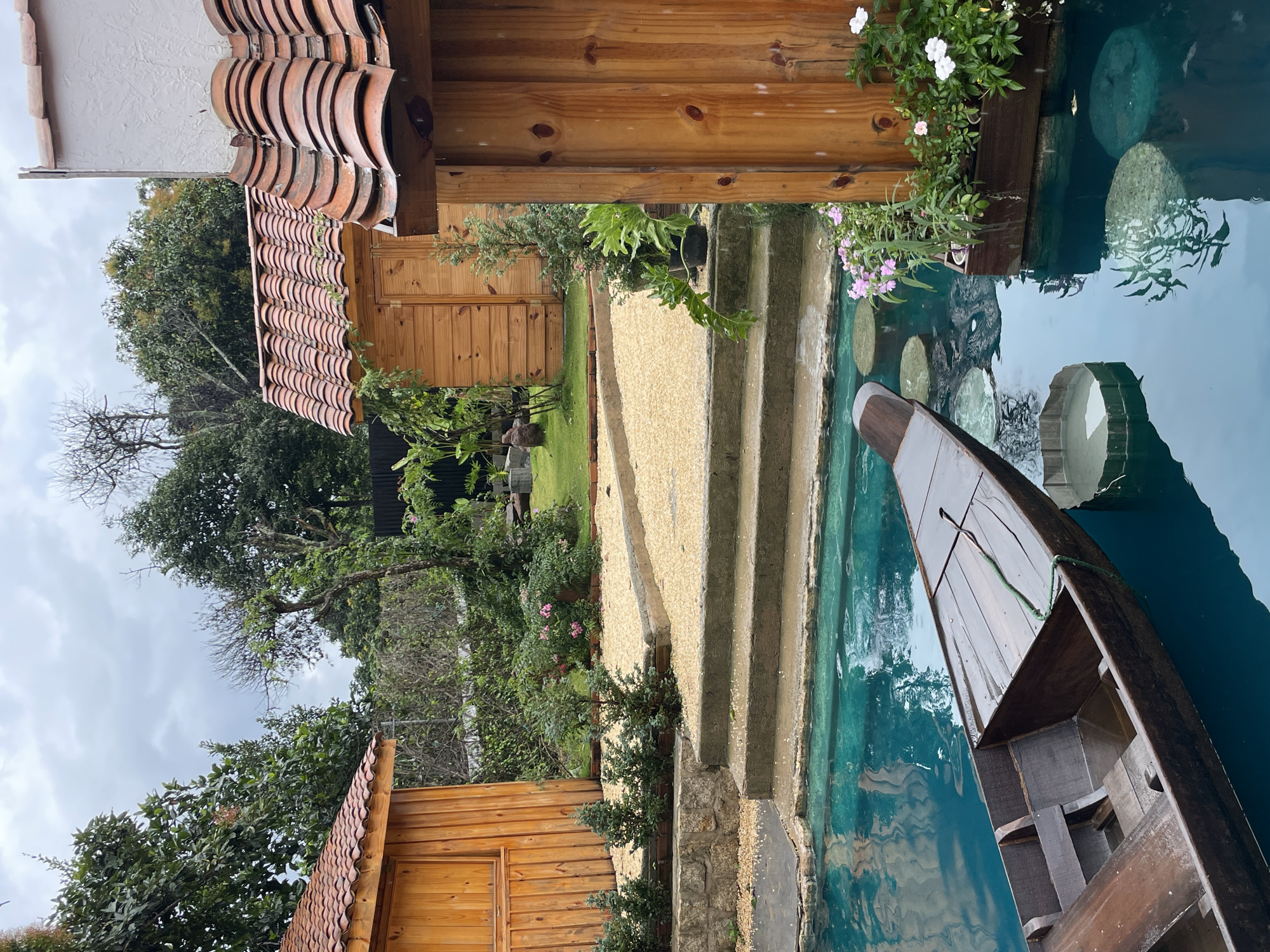 The image depicts an outdoor scene with a body of turquoise water in the foreground. A wooden boat is partially submerged in the water, pointing towards the left side of the frame. In the middle ground, there are concrete steps leading up to a gravel-covered area. Several small, conical evergreen trees are planted along a low stone wall. Beyond the steps, two wooden structures with red tiled roofs are visible. The structure on the left is a simple wooden building, while the structure on the right appears to be a smaller shed or outbuilding. Lush green trees and bushes surround the area, and the sky is overcast. To the right of the steps, a wooden planter box filled with pink and white flowers floats in the water. This scene appears to be a tranquil, possibly resort-like setting. The provided context indicates this location is in Thôn 3, Vietnam.