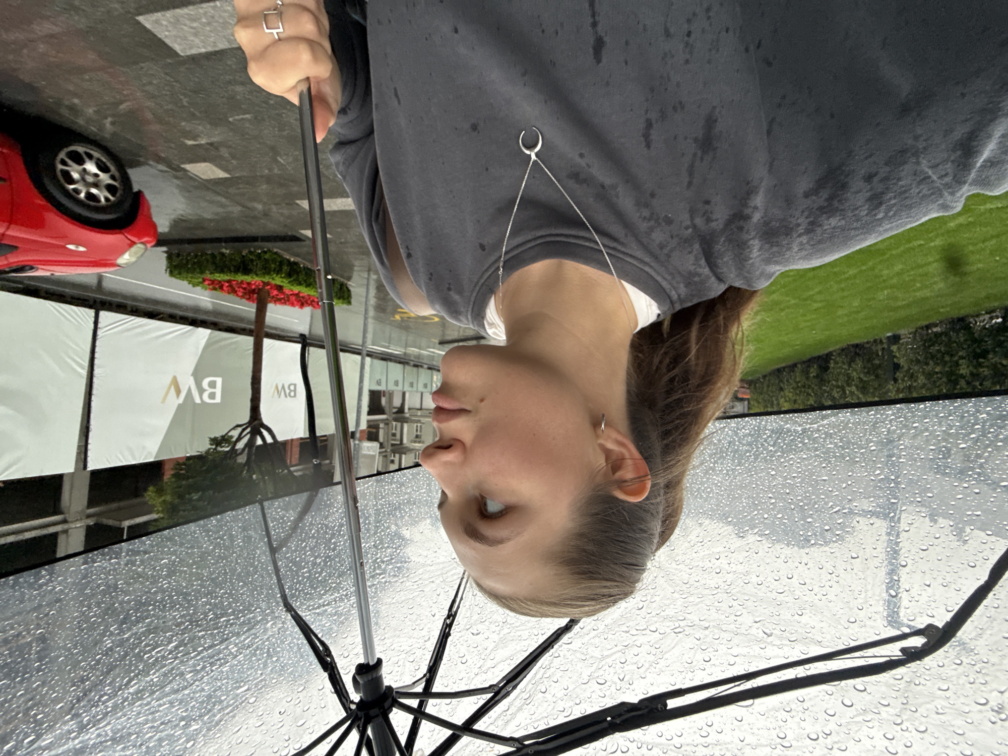 A young woman with brown hair pulled back in a ponytail is holding a clear umbrella over her head while standing outdoors. She is wearing a dark grey sweatshirt and a silver necklace with a crescent moon pendant. She is looking to her right, away from the camera. The umbrella is speckled with raindrops. In the background, there is a green lawn, a tree, a red car, and buildings under construction. The ground is wet, indicating it is raining. This scene is likely located in the city of Belgrade, Serbia, near a construction site, possibly in the Savamala district given the modern architecture and construction visible.