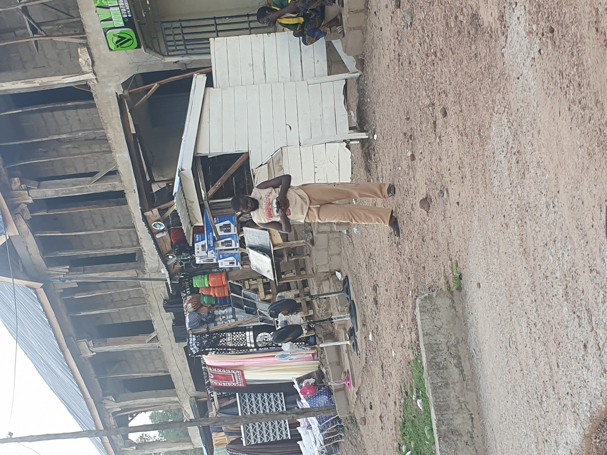 The image captures a street scene in Lokoja, Nigeria. A man stands in front of a shop, his hands clasped near his waist. The shop displays electronics, clothing, and household items. A second person sits near a white wooden structure beside the shop. The ground is dusty and gravelly. The building has a metal roof and concrete supports.