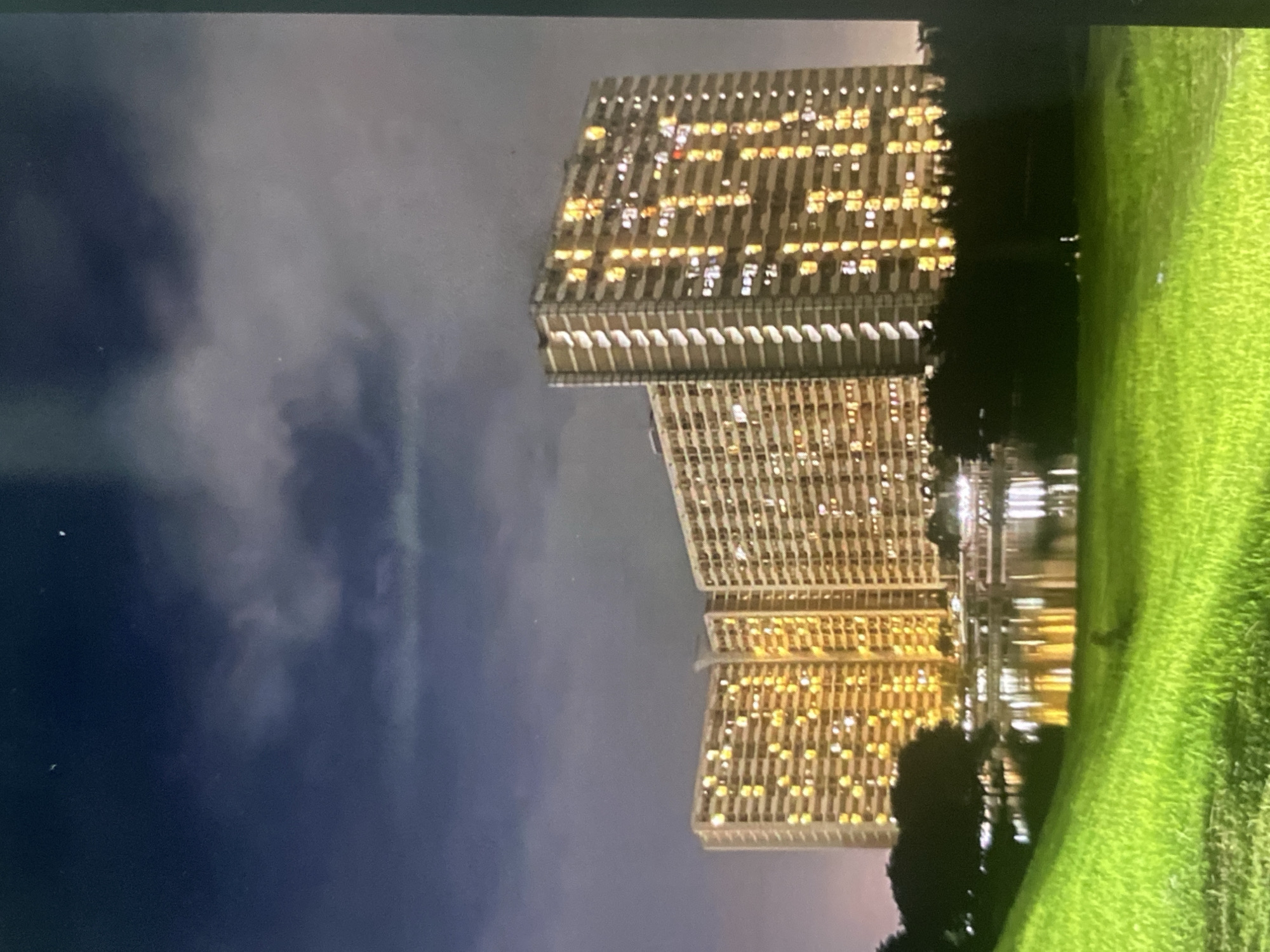 This is a nighttime photograph of three illuminated apartment buildings in the city of Tebet, Indonesia. The buildings are tall, rectangular structures with many windows, most of which are lit with warm, yellow light, suggesting occupancy. The buildings are clustered together, with the rightmost building being the tallest. In front of the buildings is a body of water, likely a lake or pond, which reflects the lights from the buildings. The water is surrounded by dark trees and foliage. In the foreground, there is a gently sloping grassy hill, illuminated by an unseen light source, giving it a vibrant green color. The sky above is dark and cloudy, with a few faint stars visible. The overall impression is of a residential area at night, with the lights from the apartments creating a warm and inviting glow against the dark sky and landscape.