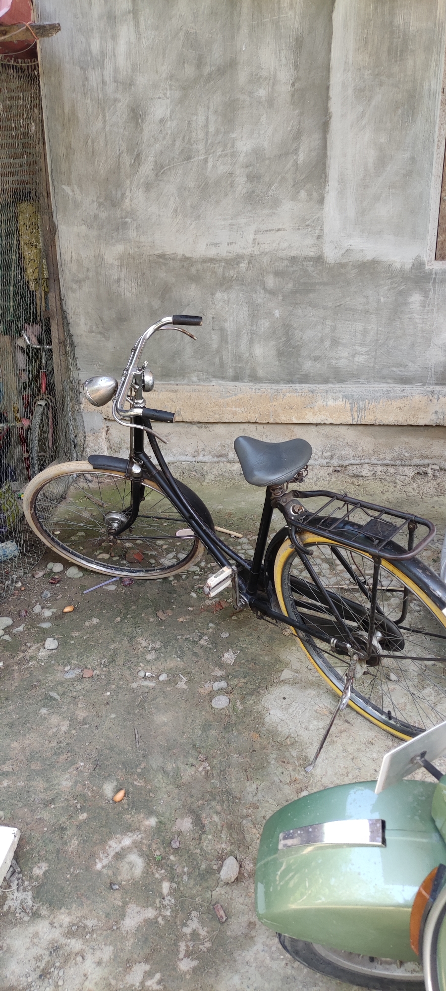 An old, black bicycle is leaning against a rough, grey concrete wall. The bicycle has a silver headlight, black handlebars, a grey seat, and a black rear rack. Its tires are yellow. To the left of the bicycle, there is a wire mesh fence with various items behind it, including other bicycles. The ground is composed of dirt and small stones. In the foreground, the rear of a light green scooter is visible. The scene appears to be outdoors, possibly in a yard or alleyway, with the general environment suggesting a possibly residential area in Payakumbuh, Indonesia.