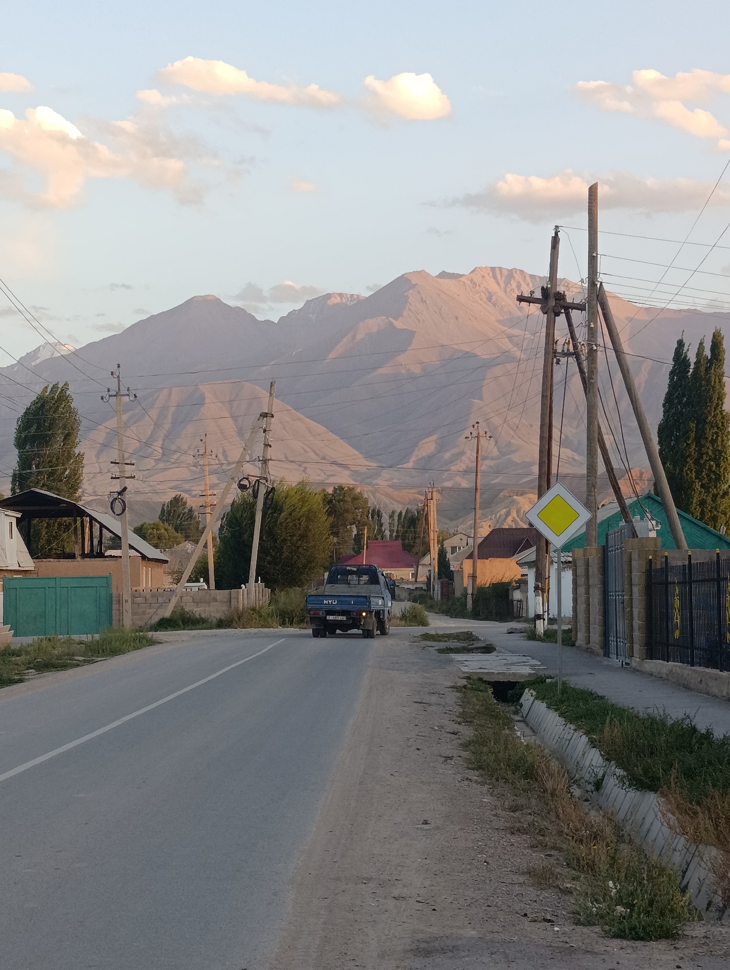 The image depicts a street scene in Kochkor, Kyrgyzstan, with a backdrop of majestic mountains. A paved road runs through the center of the frame, with a white line marking the lane. A blue truck is visible driving on the road. To the left of the road are houses and a green metal gate. To the right, a sidewalk is lined with a metal fence and a yellow diamond-shaped road sign. Telephone poles and power lines are scattered throughout the scene, with lush green trees interspersed amongst the buildings. The sky above is a soft blue, with scattered clouds tinged with the warm hues of either sunrise or sunset, casting a gentle light on the mountains.