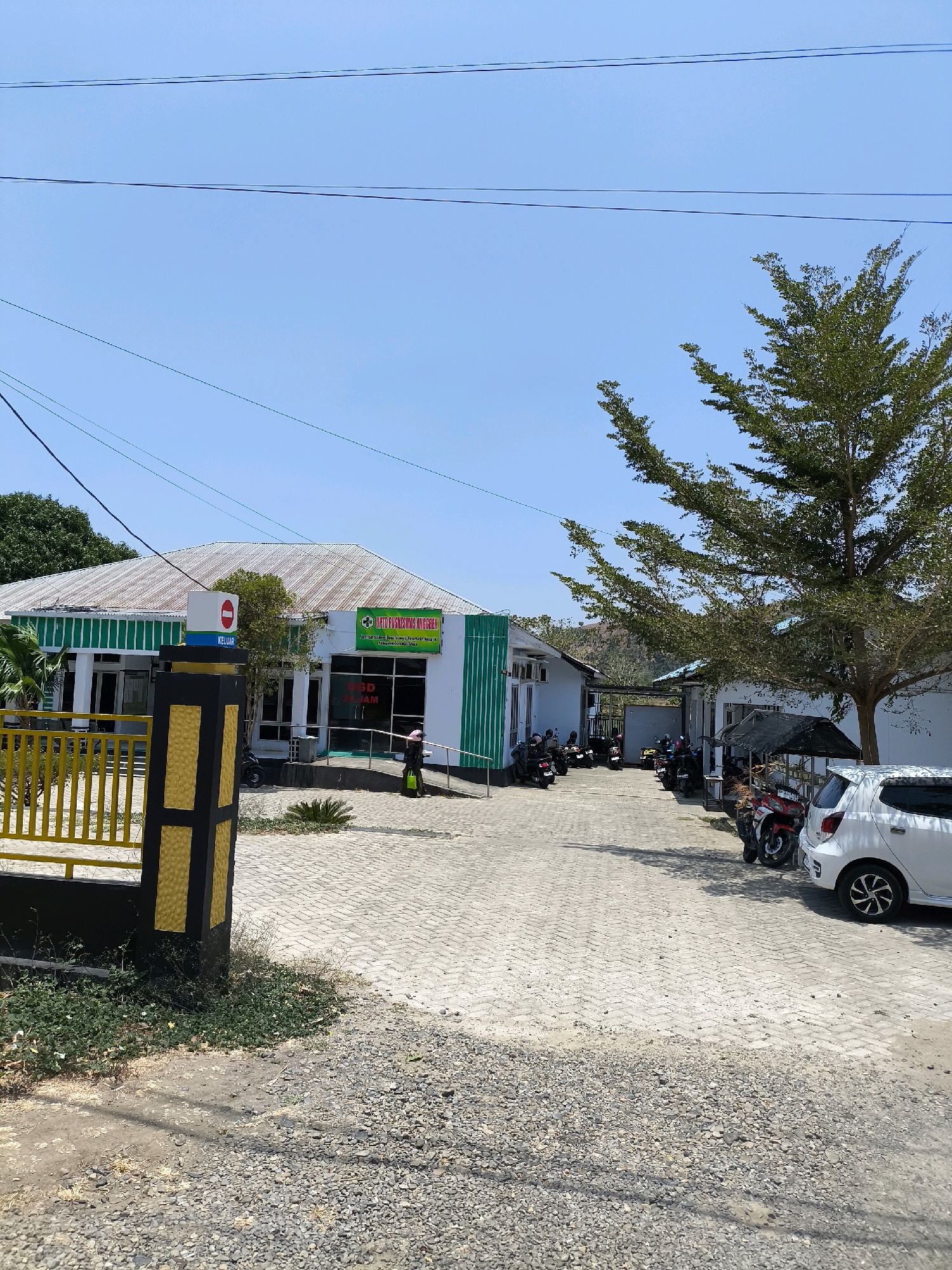 The image depicts the exterior of a healthcare facility, identified by signage as "PUSKESMAS ANGGREK" (Anggrek Community Health Center). The building, painted white with green accents, has a corrugated metal roof. A sign with a red circle and a horizontal bar, indicating "STOP" or "DO NOT ENTER," is visible near the entrance, along with a blue sign that reads "KELUAR" (EXIT). In front of the building, a paved walkway leads to an entrance with a ramp. Several motorcycles are parked in the foreground and to the side of the building. A white car is parked in the lower right portion of the image. A person wearing a pink head covering and a green garment is walking near the ramp. The environment appears to be an outdoor area with a clear blue sky, a large tree with sparse foliage, and some vegetation. The scene suggests a functional, everyday operation of a community health center in a sunny, possibly tropical, location in Indonesia.