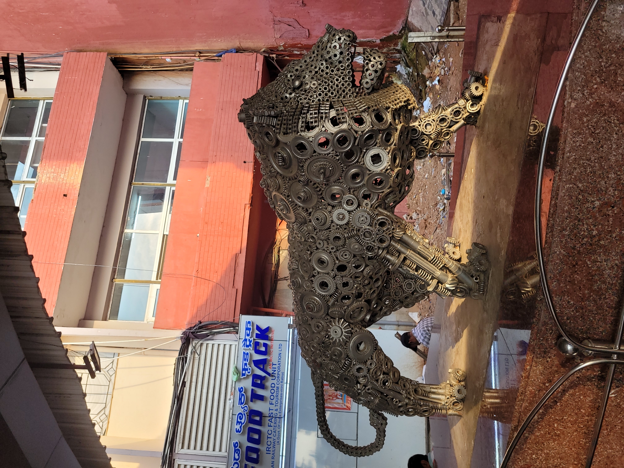 The image displays an outdoor urban scene featuring a prominent sculpture of a predatory animal, likely a lion, crafted entirely from intricate metallic components like gears, bearings, chains, and other mechanical parts. The sculpture is large and detailed, depicted in a dynamic pose, possibly mid-stride or pounce, with its tail curving around its body.
In the background, a multi-story building with a reddish-orange facade and multiple windows is visible. Below, a stall or shop entrance features a prominent sign. The visible text on the sign reads: "FOOD TRACK," "AN IRCTC INITIATIVE," "IRCTC FAST FOOD UNIT," and "(A Govt. Of India Enterprises-mini Ratna) AN RAILWAY CATERING & TOURISM CORPORATION LTD." The sign also includes text in Kannada script, "ಫುಡ್ ಟ್ರ್ಯಾಕ್."
Two individuals are partially visible in the background; one is obscured by the sculpture's tail, and another is seated further back to the right. The ground immediately surrounding the sculpture is uneven, with visible debris and litter, while the foreground shows a clean, reddish-brown speckled tiled floor with a curved metal railing. The bright sunlight indicates it is daytime, casting noticeable shadows from the left, suggesting either morning or afternoon. The scene depicts a public area, likely a commercial or transit zone given the IRCTC affiliation.