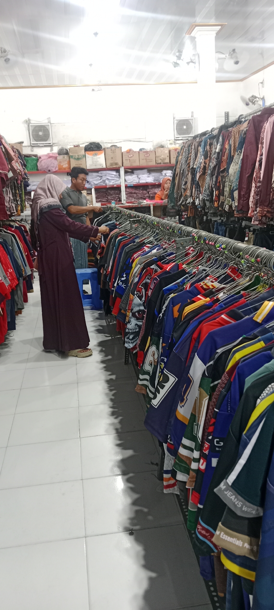 In Rengel, Indonesia, a retail store displays clothing on racks. A woman wearing a maroon hijab and dress is examining shirts on a rack, with a man in a grey polo shirt standing behind her. Another woman in an orange top is visible in the background. The store features white tiled flooring, fluorescent lighting, and shelving stocked with boxes and clothing.