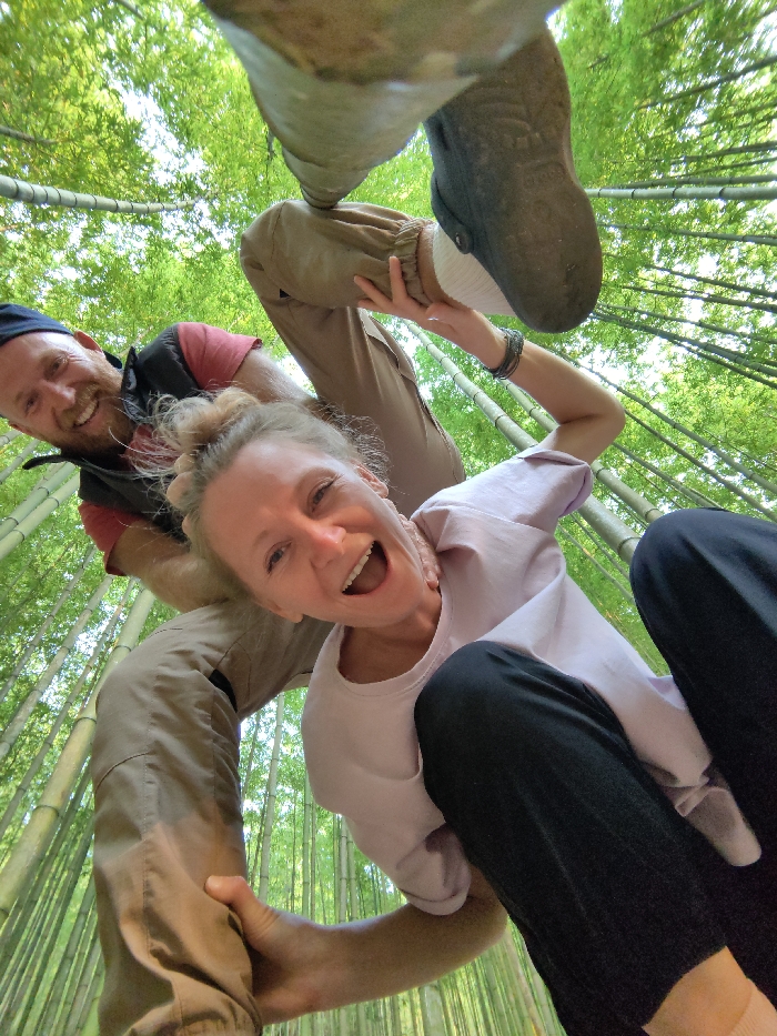 A wide-angle, low-perspective photograph depicts two individuals in a dynamic pose within a dense bamboo forest in Mù Cang Chải, Vietnam. The environment is characterized by numerous slender green bamboo stalks and foliage extending upwards, filling the background. A male subject, visible on the left, wears a blue cap, red t-shirt, dark vest, and light brown pants. He has a red beard and smiles directly at the camera. His right leg is bent, with his dark-shoed foot elevated prominently at the top center of the frame. A female subject, positioned closer and to the right, has blonde hair in a bun, wears a light pink t-shirt and dark pants. She smiles widely with an open mouth, looking at the camera. Her left hand or arm is observed grasping the male's lower left leg. Both individuals are engaged in a posed interaction, with the female appearing to be supported or partially lifted by the male.