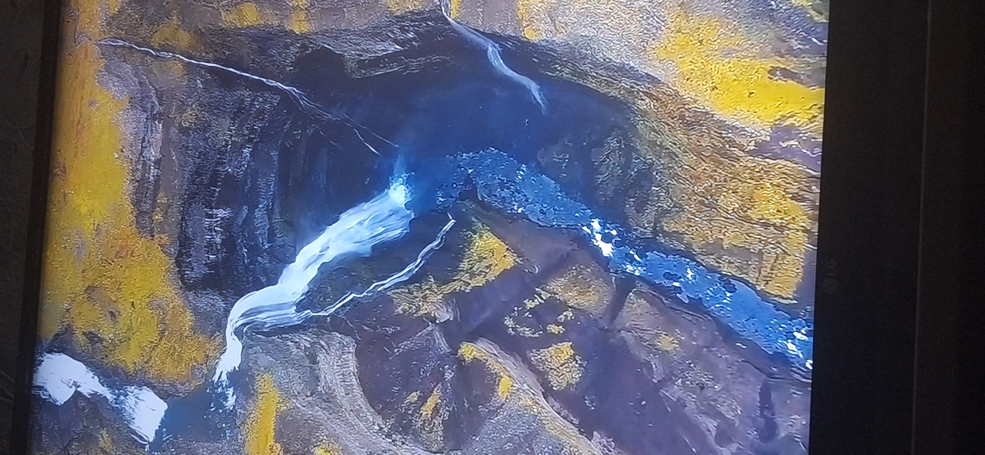 The image is a photograph of a television screen displaying an aerial view of a waterfall cascading into a gorge. The surrounding landscape is rugged and appears to be covered in a mixture of dark rock and yellow vegetation. The water is a vibrant blue, creating a striking contrast with the earthy tones of the terrain. The image does not depict any elements that would indicate a location within the city of Bila Tserkva, Ukraine, nor does it show any people or specific man-made objects other than the television frame and the LG logo at the bottom.
