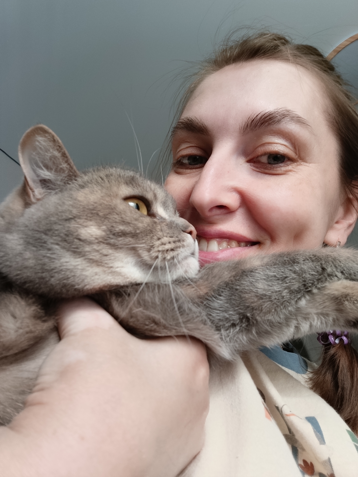 A woman with fair skin, light brown hair partially pulled back, and a bright smile is holding a grey cat with yellow eyes. The woman's face is slightly angled toward the camera, and her smile reveals her upper teeth. The cat is held close to her face, with its head turned to the left, away from the camera. The woman's hand is visible, holding the cat around its neck. Part of her shirt is also visible, showing a pattern of beige and light blue colors with illustrations on it. The background appears to be a plain grey wall. The location is the city of Chaikovsky, Russia.