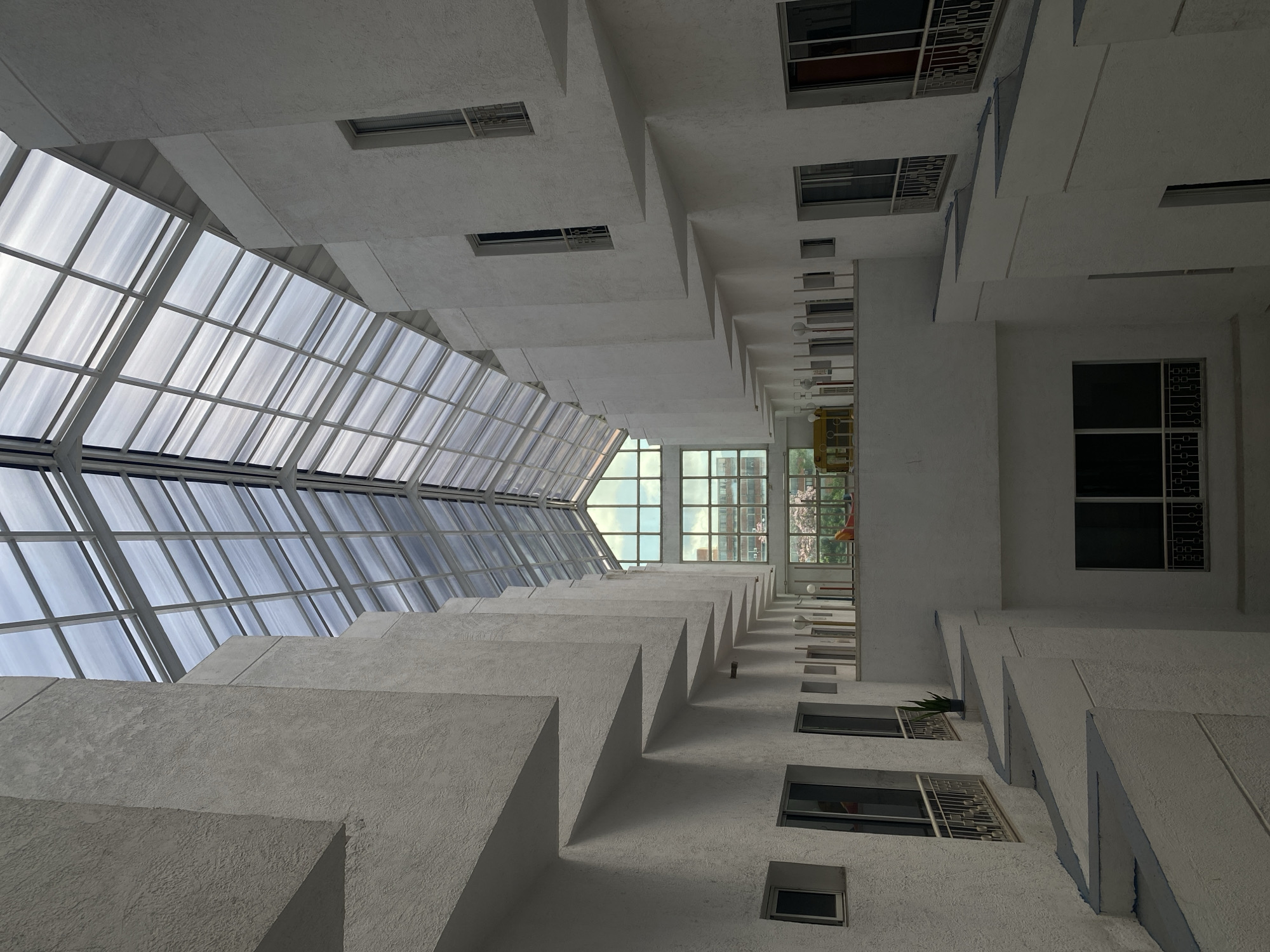 The image depicts an upward view of a building facade, potentially located in Naguanagua, Venezuela. The structure exhibits a geometric design with protruding blocks and inset windows. A glass ceiling covers a portion of the space, allowing natural light to enter. The building's exterior is predominantly white or light-colored. Windows of various sizes are visible, some with metal grilles. The perspective is from below, looking up towards the sky.