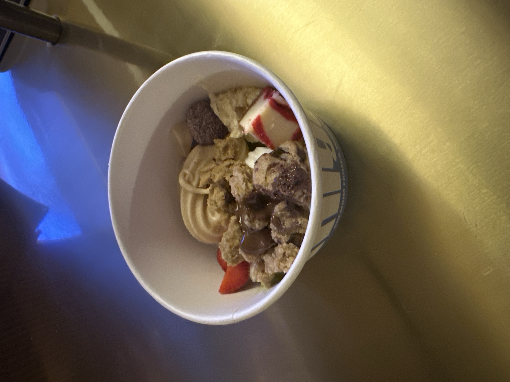 The image presents an overhead view of a white paper cup filled with a dessert, likely frozen yogurt or ice cream, and an assortment of toppings. No people are visible.
The contents of the cup include a swirl of light-colored frozen yogurt or ice cream, generously topped with various items. Among the toppings are chunks of crumbled cookies or cake, some of which are drizzled with a dark sauce, possibly chocolate. A prominent piece of white fudge or candy with red stripes is visible towards the back of the cup. Other visible toppings include a reddish-brown, textured chunk, possibly a brownie bite, and smaller white pieces that resemble marshmallows. A fresh, red strawberry slice is also nestled towards the bottom left of the cup's contents.
The cup rests on a reflective surface, which exhibits a golden-yellow hue on the right side and transitions to a deep blue on the left, indicating a strong blue light source reflection. A dark, cylindrical object is partially visible in the upper left corner of the background. The exterior of the cup shows fragmented text in dark blue, with partial words like "FROZEN" and "PREMIUM" discernible, suggesting the type of product. The lighting is artificial and uneven, creating distinct reflections and shadows. The scene captures a close-up of a prepared dessert. The location context for this media file is Mengwi, Indonesia.