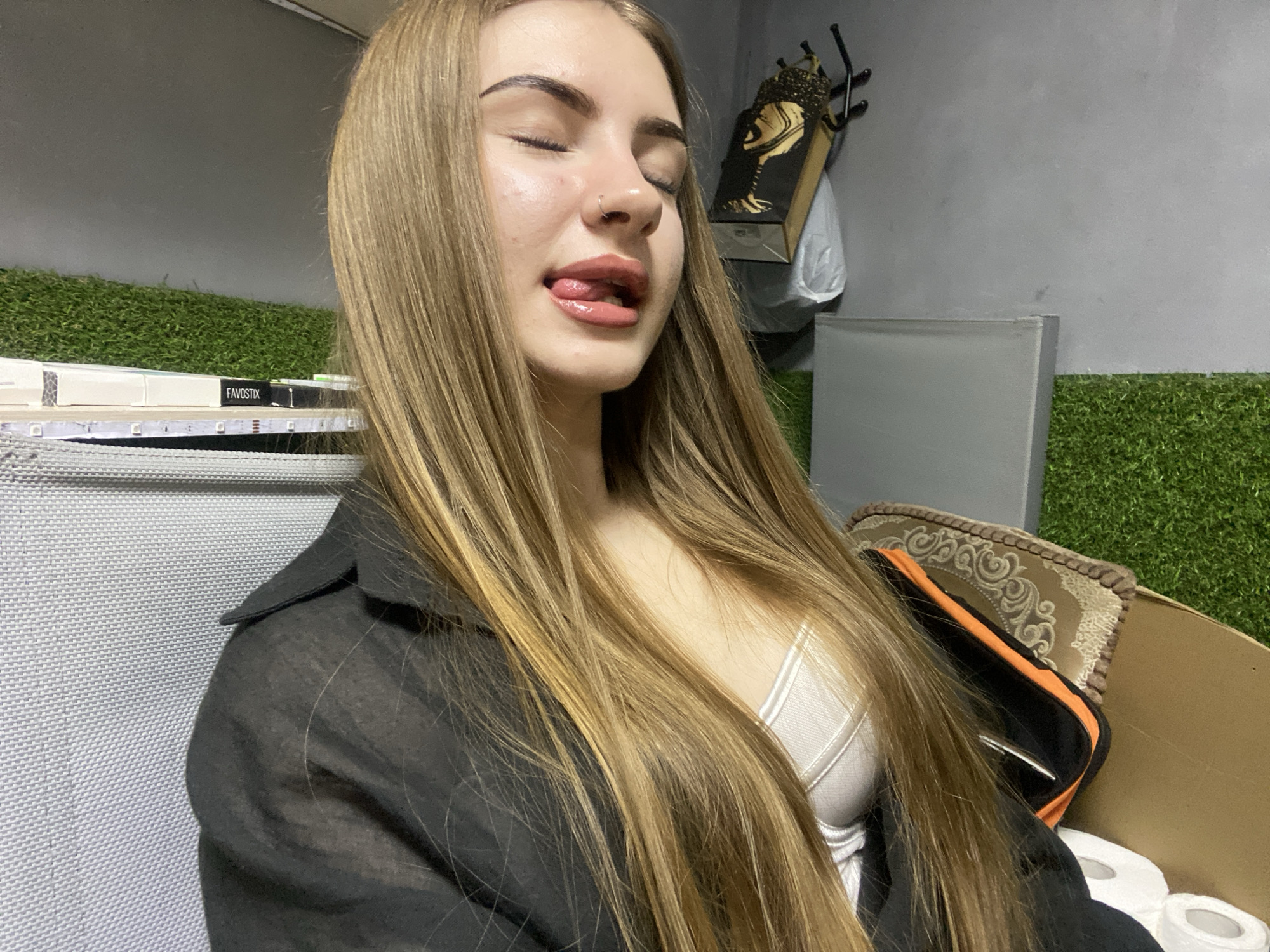 A young Caucasian woman with long blonde hair and makeup is pictured indoors, likely in a residential setting in Dyakovo, Russia. She is wearing a black unbuttoned shirt over a white top. Her eyes are closed, and her tongue is sticking out, giving a playful or suggestive expression. In the background, artificial green turf is visible along the bottom of the wall. To the right of the woman, there's a cardboard box containing rolls of toilet paper and an orange and black pouch resting on a decorative pillow. A wall-mounted decorative item with a gold abstract design is also visible on the grey wall. The lighting appears to be artificial and slightly dim.