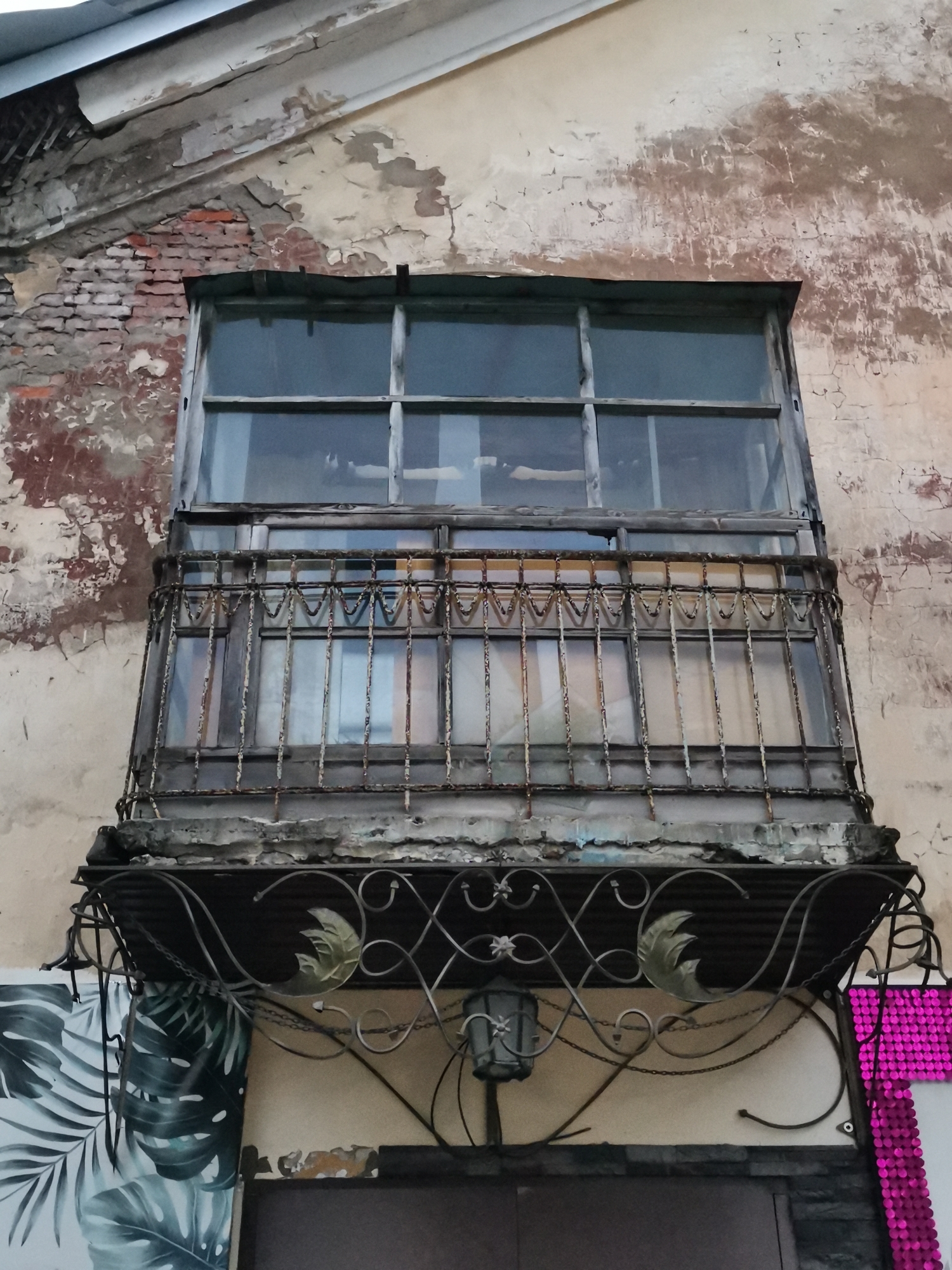 An outdoor, low-angle shot captures a weathered facade with a dilapidated balcony in Alexandrovsk, Russia. The building's exterior exhibits peeling paint and exposed brickwork, suggesting age and neglect. The central focus is a small, enclosed balcony with a glass window divided into panes by wooden dividers. A rusted metal railing with an ornate pattern runs across the front of the balcony. Below the balcony, a decorative metal canopy with leaf motifs hangs above an entrance. To the left and right of the entrance, portions of large, stylized tropical leaf murals are visible. A vintage-style lantern is suspended from the underside of the canopy. The overall scene conveys a sense of historical decay and artistic embellishment.