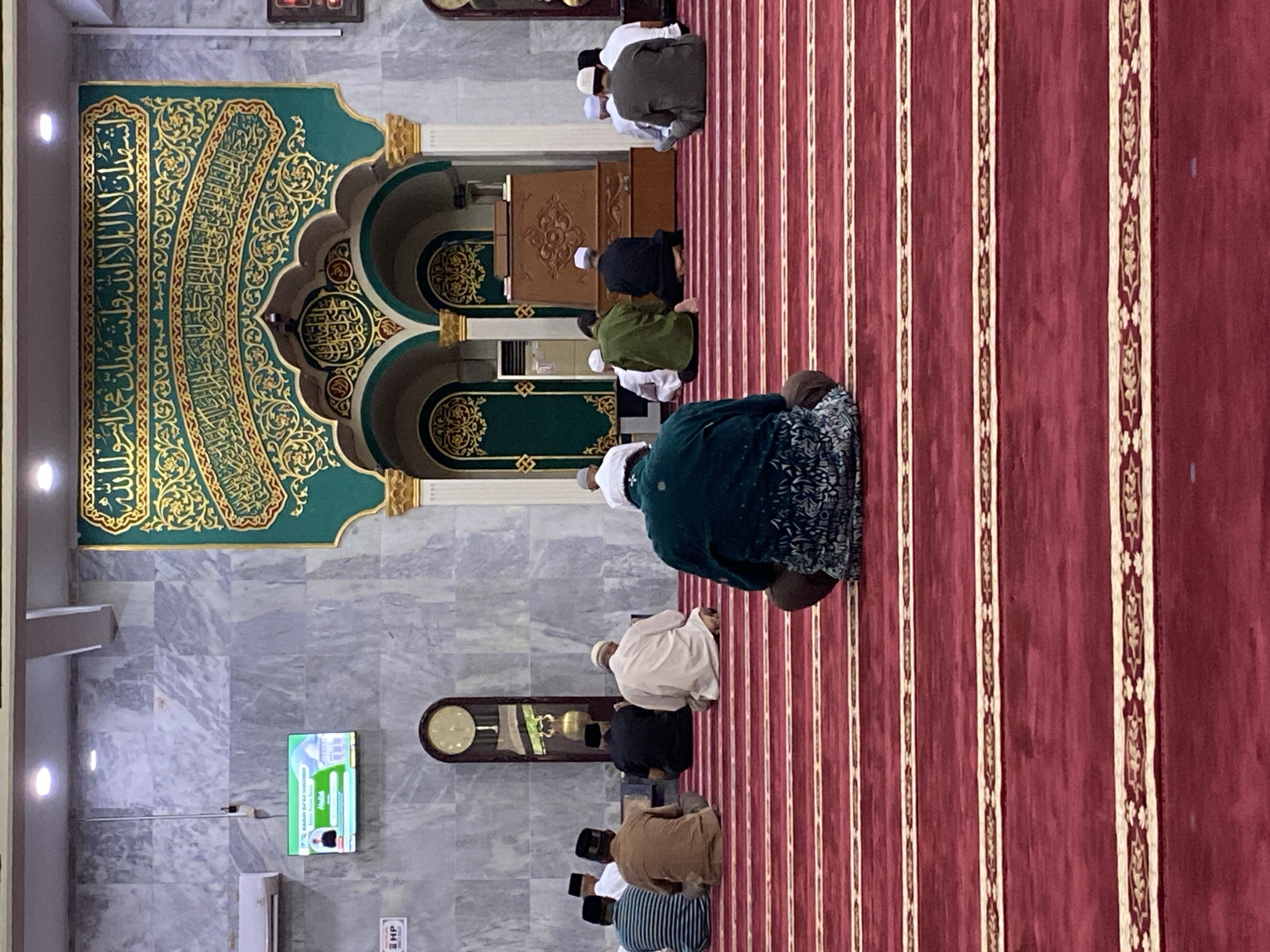 The image shows the interior of a mosque, likely in Lhokseumawe, Indonesia, with several men sitting on a red carpeted floor, facing a green and gold mihrab. The mihrab is elaborately decorated with Arabic calligraphy and intricate patterns. In the foreground, a man in a dark green tunic with a patterned design is seated with his back to the camera. Behind him, other men are seated in rows, some wearing white caps and traditional Islamic attire. To the left, a large grandfather clock stands against a marble-patterned wall. A television screen displaying an image of a mosque and text is mounted on the wall above the clock. The mosque has high ceilings with spotlights and decorative gold accents on the pillars. The overall atmosphere appears serene and devotional.
