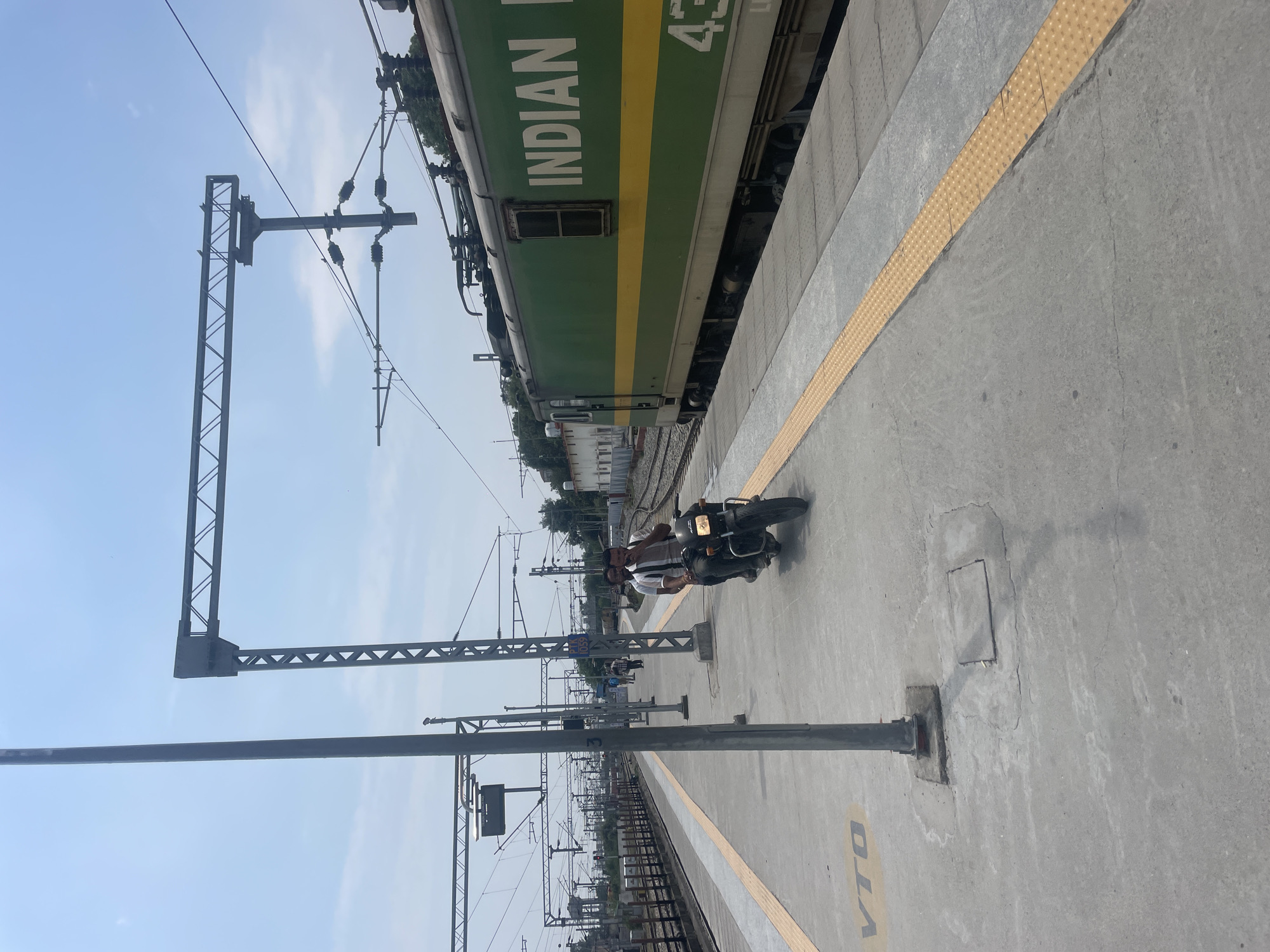 The image shows a scene at a railway station platform in Pathankot, India. A green and yellow Indian Railways electric locomotive is parked on the tracks. Two men are riding a motorcycle on the platform. In the foreground, there is a concrete platform with yellow tactile paving along the edge. Several poles and overhead electrical equipment for the train are visible. The sky is clear blue with some clouds. The overall environment appears to be an active railway station.
