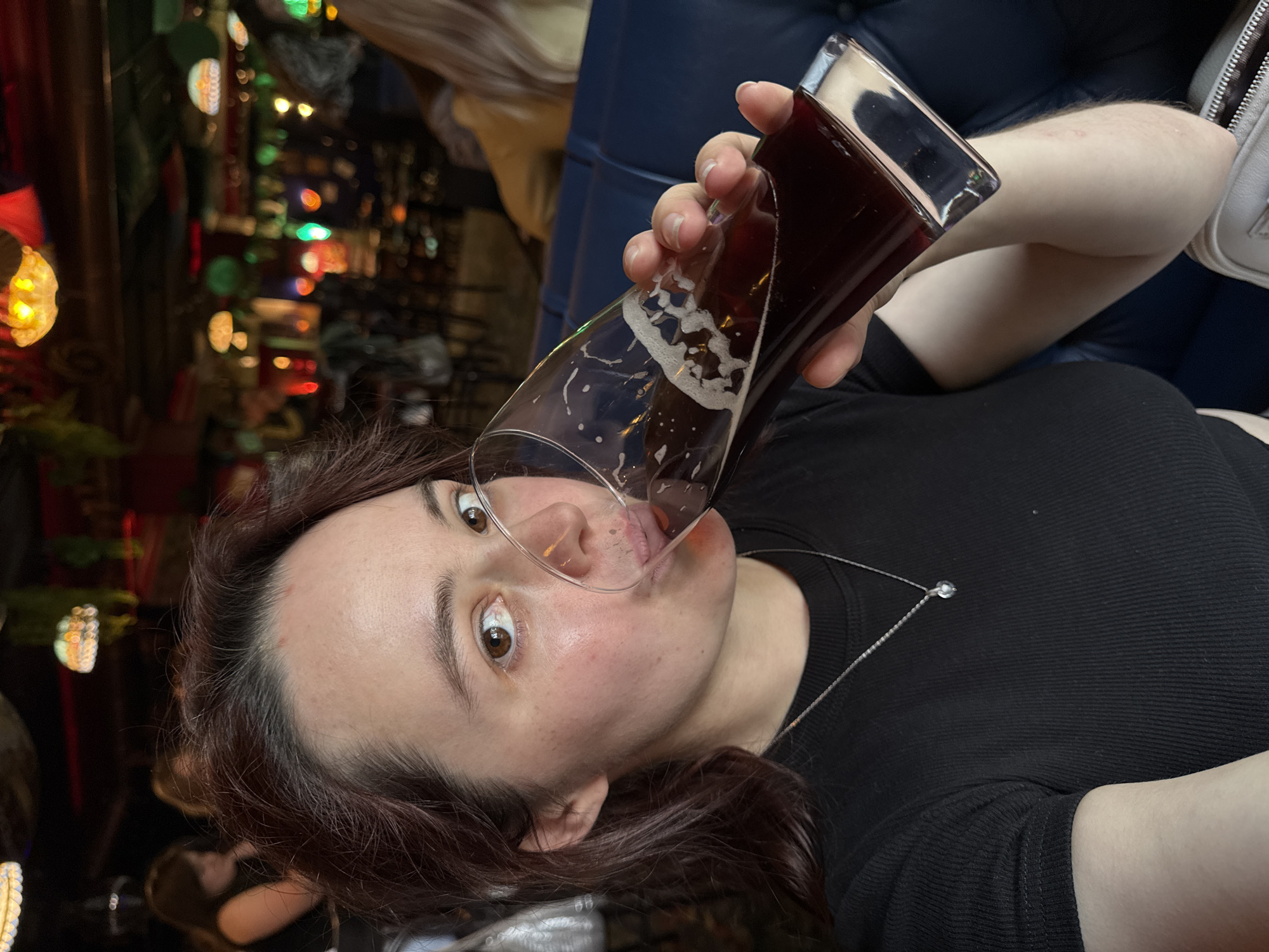 A woman is captured in a close-up shot, holding a glass of dark liquid, possibly beer, to her lips. She is wearing a black ribbed t-shirt and a delicate silver necklace with a pendant. Her right hand, with painted nails, grips the glass, while her left arm is resting on a dark blue upholstered surface. The background is blurred, revealing a dimly lit indoor environment with a warm, inviting ambiance, suggesting a bar or restaurant. The lighting, with a mix of warm yellow and red hues from overhead fixtures, contributes to the cozy atmosphere. There are other patrons in the background, also blurred, with one individual on the left wearing a dark top and another on the right with blonde hair. The image appears to have been taken in Izhevsk, Russia.