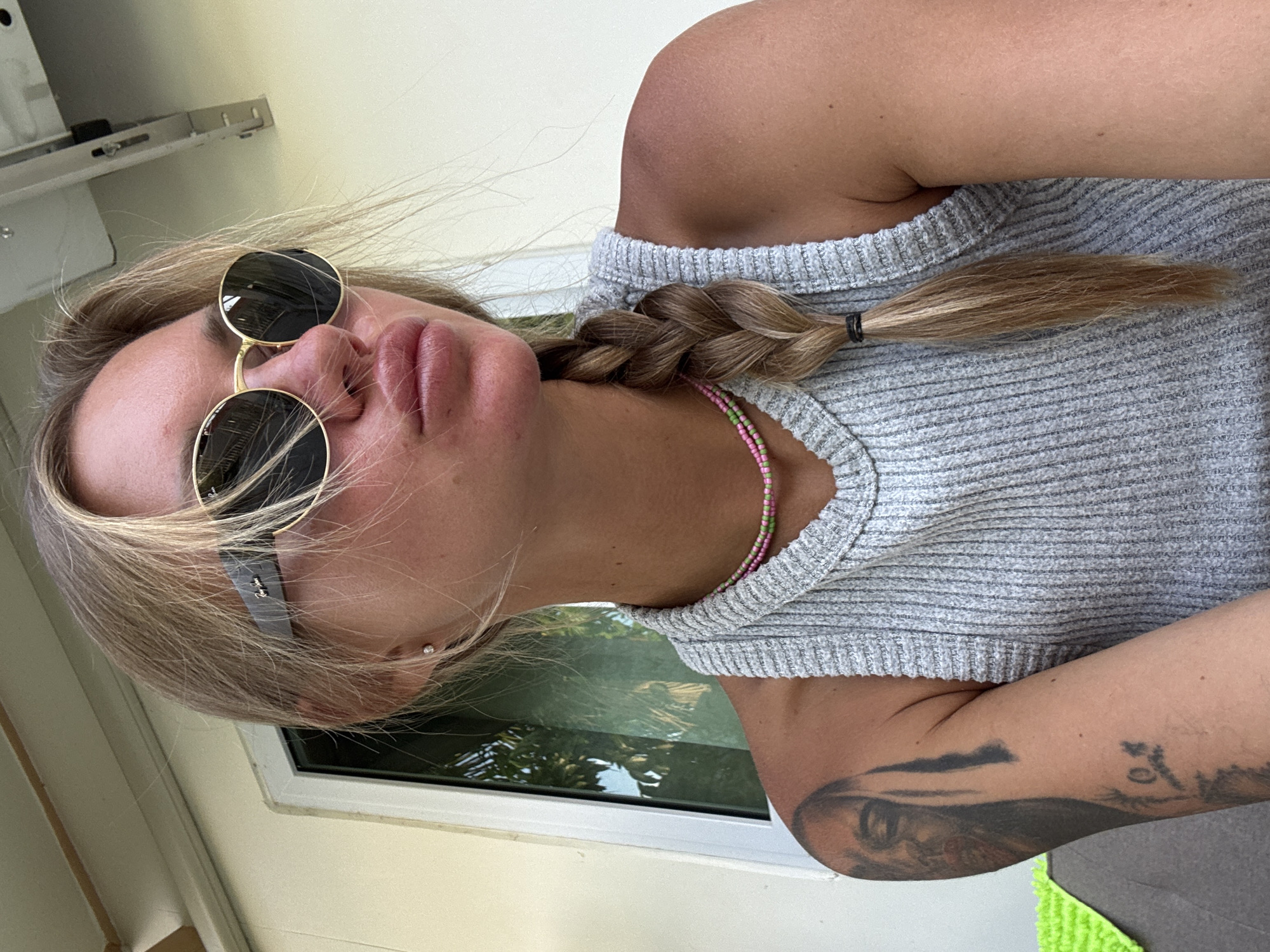 A blonde female is depicted in a close-up, chest-up portrait in Choeng Thale, Thailand. She is positioned slightly to the right of center, looking towards the upper right of the frame with her lips puckered. She wears dark, round sunglasses with gold frames, a light gray ribbed sleeveless top, and two beaded necklaces (one pink, one green) around her neck. Her blonde hair is styled in a side braid, secured with a black elastic band, resting on her right side. A small, clear stud earring is visible in her left earlobe. A prominent tattoo featuring a face and additional symbols is visible on her left upper arm. The background consists of a light beige wall, a white window frame reflecting green foliage, and a section of a light-colored ceiling structure with a metal bracket visible above her right shoulder. A dark gray surface with a bright green textured object is partially visible to the left of her arm.