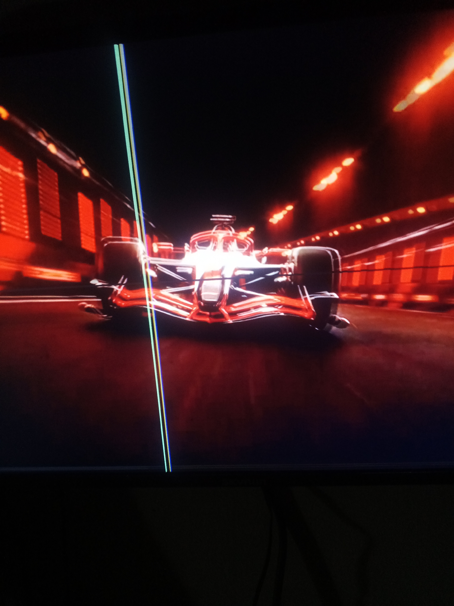 The image shows a Formula 1 race car driving down a track. The car is red and white, with a white stripe down the middle. The track is dark, with red lights on either side. There are two thin vertical lines on the left side of the image, one light green and one blue, which appear to be artifacts from the display.