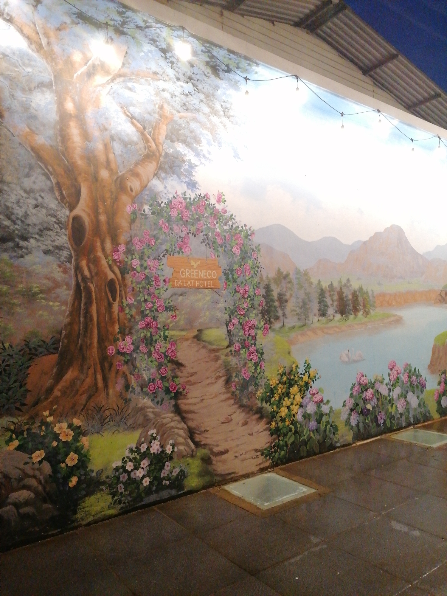 A mural is painted on an exterior wall. The painting depicts a stylized landscape scene with a large tree on the left, flowers and a path in the center, and a lake and mountains in the background. A sign in the painting reads "GREENECO DALAT HOTEL." The mural is located in the city of Da Lat, Vietnam. The foreground shows a tiled pavement with square, glass-covered features. Above the mural, a building's roofline is visible with string lights hanging below it.