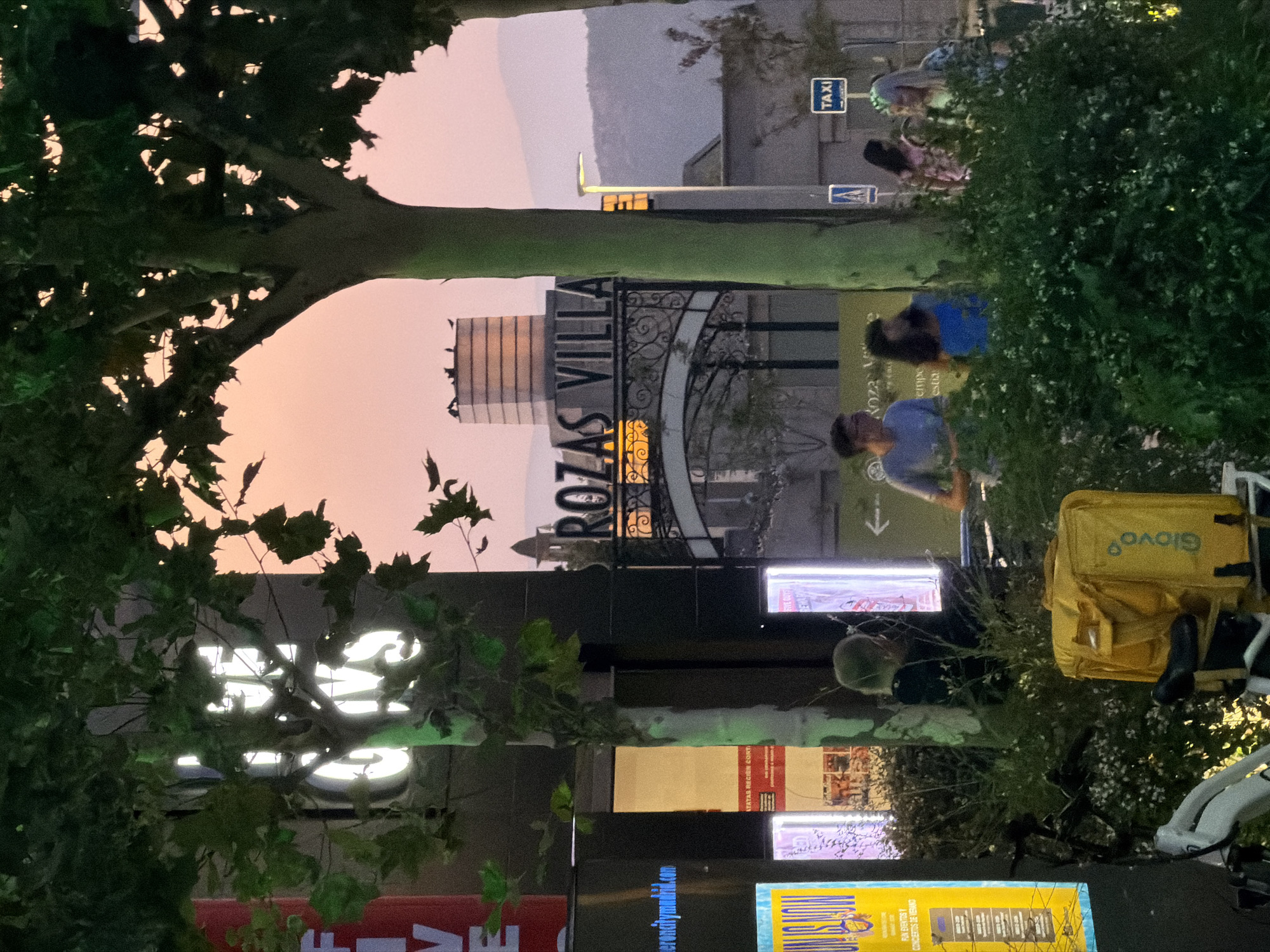 The image depicts a street scene in Las Rozas de Madrid, Spain, during twilight. In the foreground, there are trees with lush green leaves, partially obscuring the view. To the left, a bright yellow delivery bag from Glovo is attached to a bicycle. Several people are visible in the middle ground. One individual, a man wearing a blue t-shirt, stands with a woman in a blue top, and another woman in a patterned top and a woman with blonde hair in a floral top are also present. Behind them, illuminated signs are visible. The background features a building with a sign reading "ROZAS VILLA" above an ornate archway. Further in the distance, the silhouette of mountains is visible against a soft pink sky. A "TAXI" sign is also present in the middle ground. The overall impression is that of a lively evening in a commercial or entertainment district.