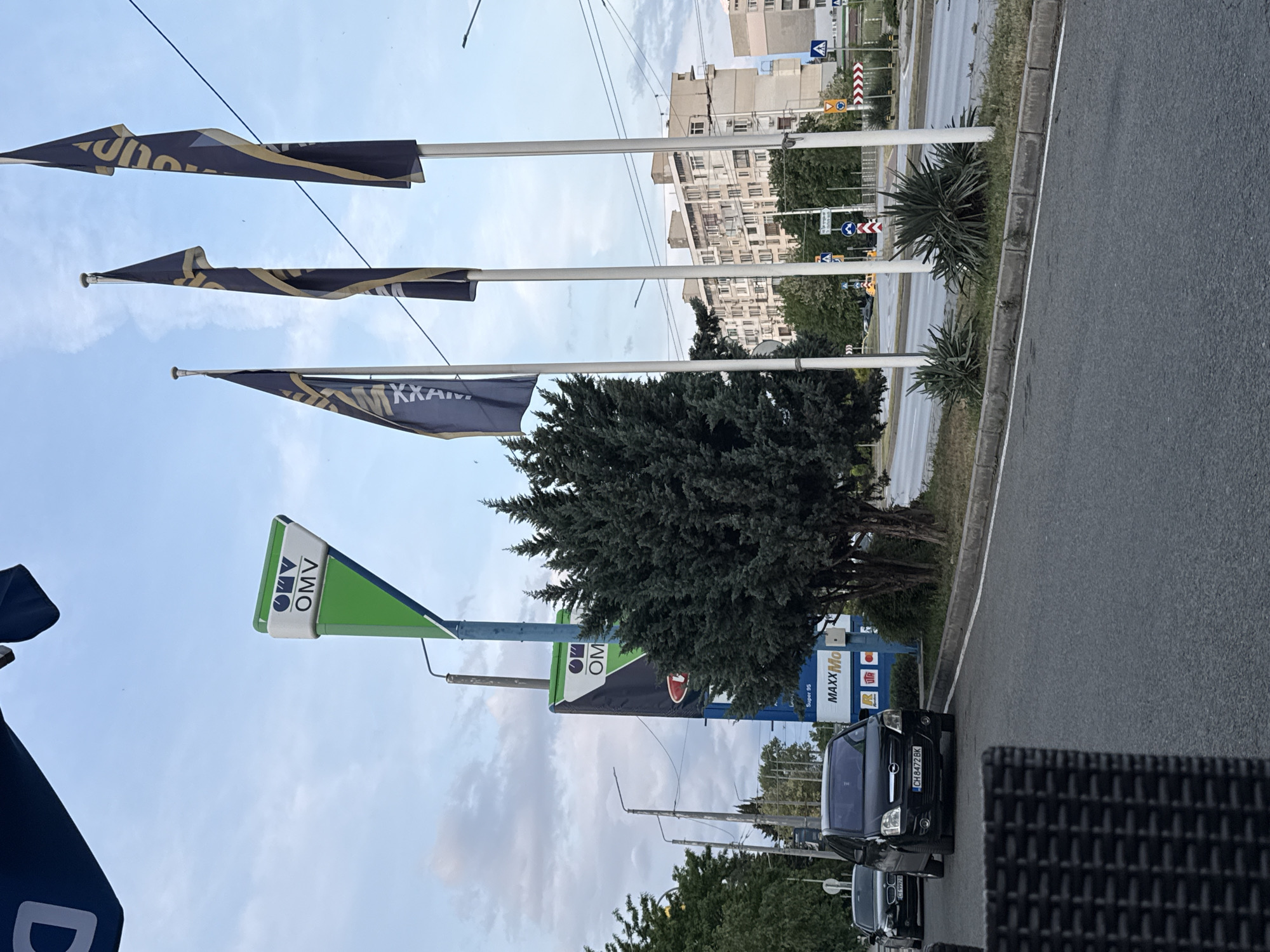 The image depicts a street scene in Stara Zagora, Bulgaria, featuring an OMV gas station with accompanying flags and signage. The street runs parallel to the gas station, with vehicles parked along the curb. Buildings are visible in the background, suggesting an urban setting. The sky is partly cloudy.