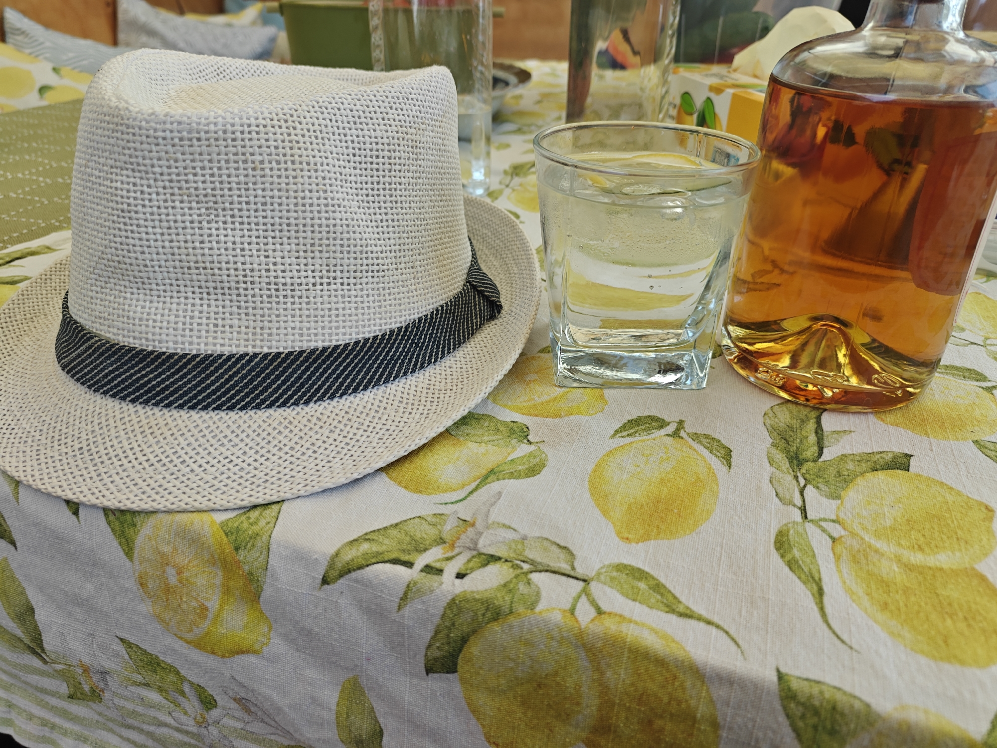In Cholpon-Ata, Kyrgyzstan, a white straw fedora hat rests on a tablecloth patterned with lemons and green leaves. Next to the hat is a short glass filled with a clear liquid and slices of lemon. To the right of the glass is a clear glass bottle containing an amber-colored liquid, likely a spirit. In the background, out of focus, are other glasses and what appears to be a tissue box with a lemon design. The scene suggests a relaxed, outdoor setting, possibly a picnic or a gathering on a sunny day.
