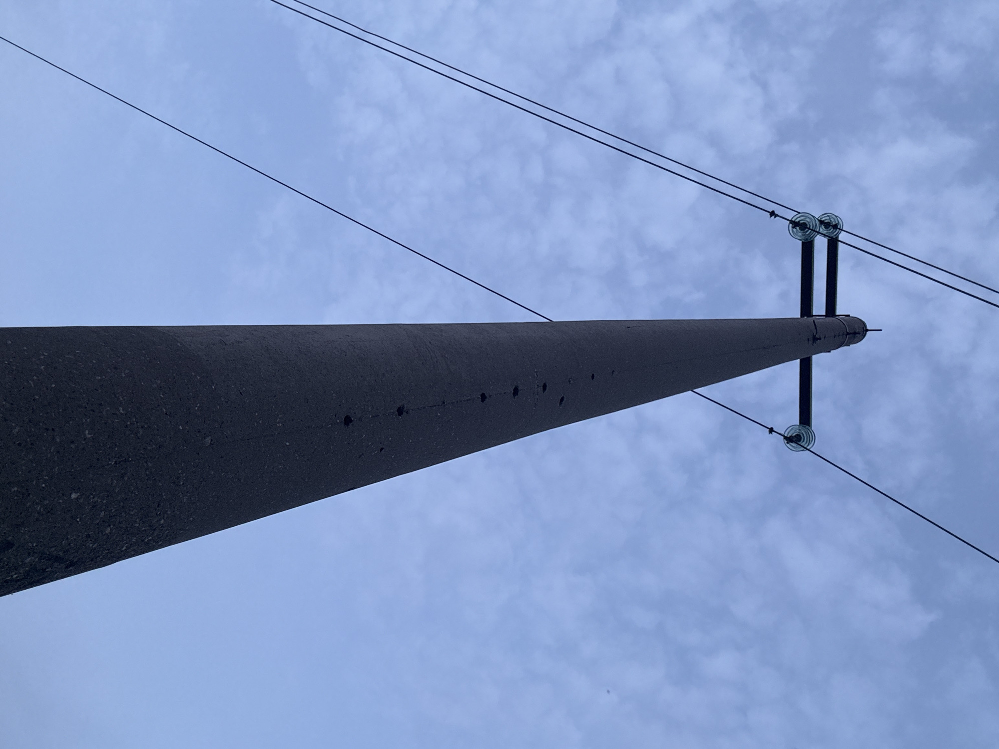 The image shows a close-up, low-angle view of a concrete utility pole against a cloudy blue sky. The pole is dark gray and appears to be made of rough concrete, with several small circular indentations visible along its length. Two dark power lines extend diagonally across the frame from the top left, supported by transparent glass insulators attached to a crossbar on the pole. The sky is overcast with scattered, thin clouds. The perspective emphasizes the height and scale of the utility pole. There are no people or animals visible in the image. The scene is likely an outdoor setting within the city of Orenburg, Russia, given the presence of a standard utility pole and overhead power lines typical of urban infrastructure.