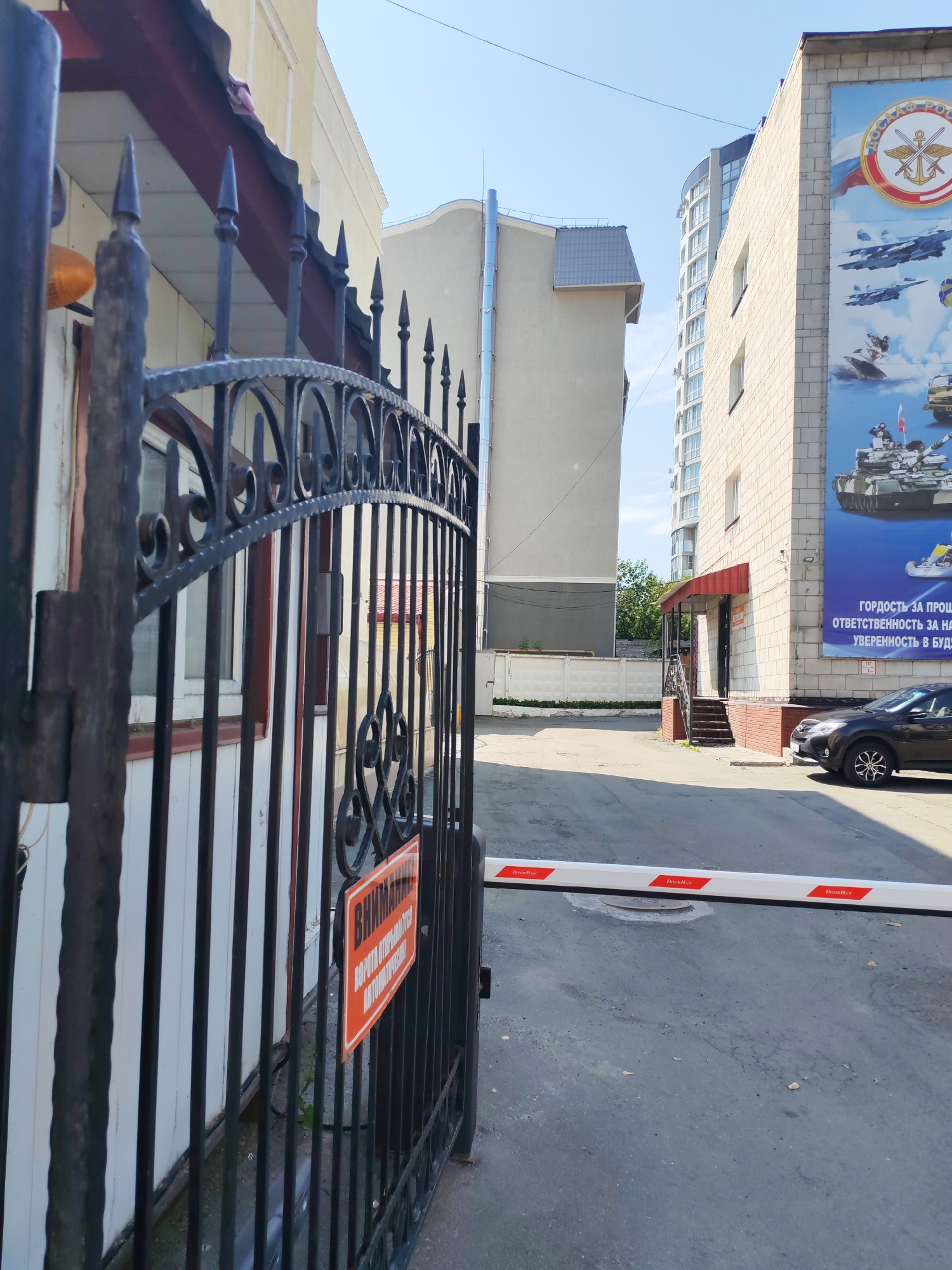 A black metal gate is in the foreground, with a sign that reads "ВНИМАНИЕ! ВОРОТА ОТКРЫВАЮТСЯ АВТОМАТИЧЕСКИ" (Attention! The gate opens automatically). Behind the gate, there is a building with white vertical siding and a dark-colored roof overhang. To the right of this building, a modern tall building with many windows is visible. Further to the right, a light-colored building with a large mural depicting military equipment, including tanks and aircraft, is present. A black sedan is parked in front of this building, and a white automatic barrier is extended across the paved area. The sky is clear and blue.