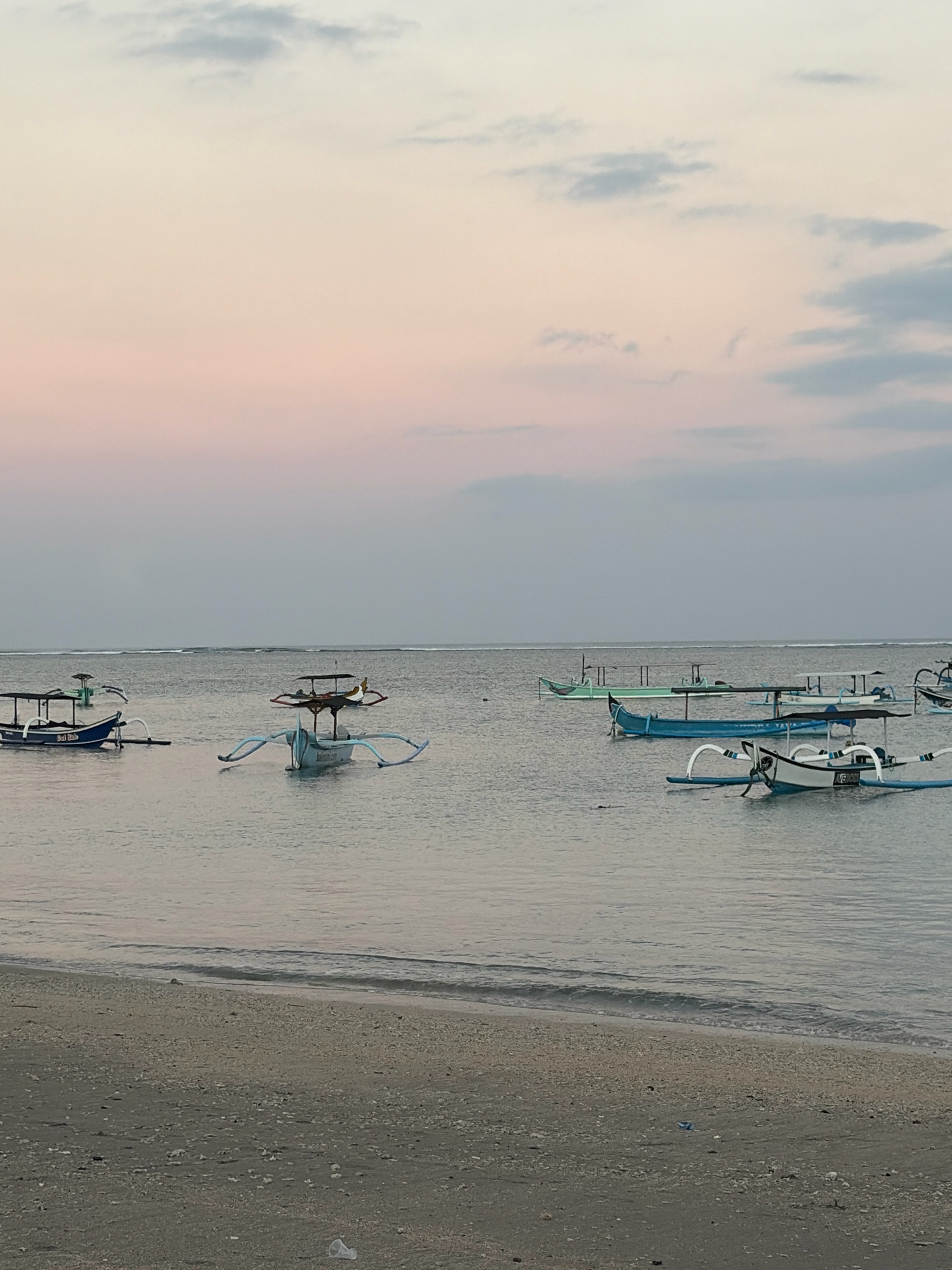 The image depicts a serene beach scene in Kuta, Indonesia, during what appears to be dusk or dawn. Several traditional outrigger boats are visible, some moored in the calm water and others closer to the shore. The sky displays a gradient of soft pastel colors, transitioning from a pale pink near the horizon to a muted gray at the top, with subtle cloud formations. The sea is tranquil, with small waves gently lapping at the sandy shore in the foreground. The beach itself is composed of fine sand with scattered shell fragments. The overall atmosphere is peaceful and contemplative, characteristic of a quiet moment by the sea.
