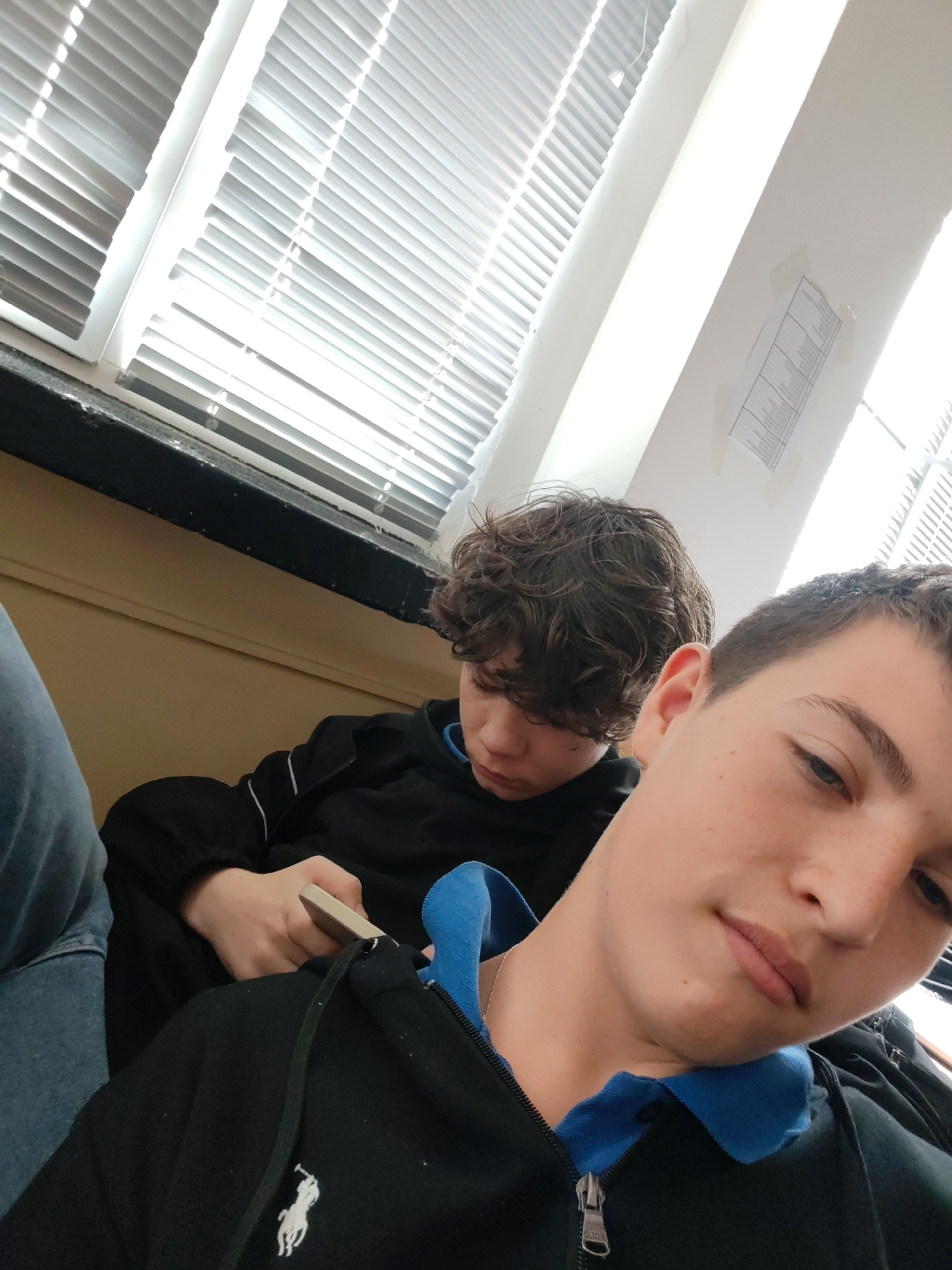This image shows two young males indoors in what appears to be a casual, brightly lit setting, likely in Burgas, Bulgaria.
In the foreground, a young male with short brown hair and light skin looks towards the camera with a neutral expression. He is wearing a black zippered hoodie with a blue collared shirt underneath, and a silver chain is visible around his neck. The hoodie features a white Ralph Lauren polo player logo on the left chest.
Behind him, slightly out of focus, sits another young male with dark, curly hair. He is wearing a black long-sleeved top, possibly a hoodie, with white stripes on the sleeves. His gaze is directed downwards, focused on a light-colored object in his hands, which resembles a smartphone. Part of a blue jeans-clad leg is visible on the far left.
The background features large windows covered by white horizontal Venetian blinds, indicating bright daylight outside. The walls are plain, with a white upper section and a light beige or yellow lower section beneath the dark-colored window sills. A faint, unreadable document or chart is taped to the white wall in the upper right. The scene suggests an interior space like a classroom, common room, or waiting area, with the individuals casually occupying their time during the day.