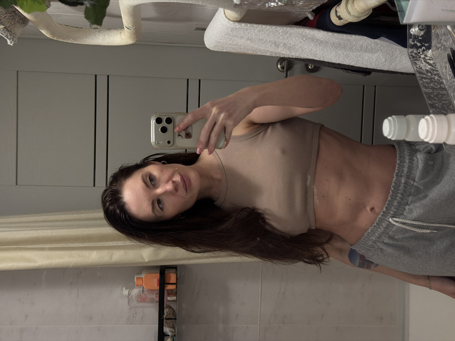 A woman with long, dark hair takes a selfie in a bathroom mirror. She wears a beige tank top and gray sweatpants, revealing her midriff. A shower curtain and bathroom products are visible in the background. The mirror reflects bathroom fixtures and toiletries. Her expression is neutral as she looks at the phone camera.